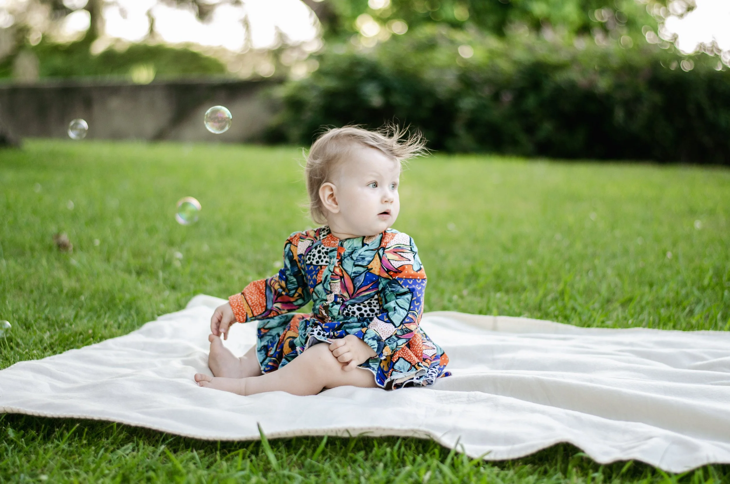 A young child with light hair wearing a colorful patterned outfit sitting on a white blanket on green grass in a park. The child looks surprised or curious as bubbles float in the air around them, with a background of blurred trees and greenery.