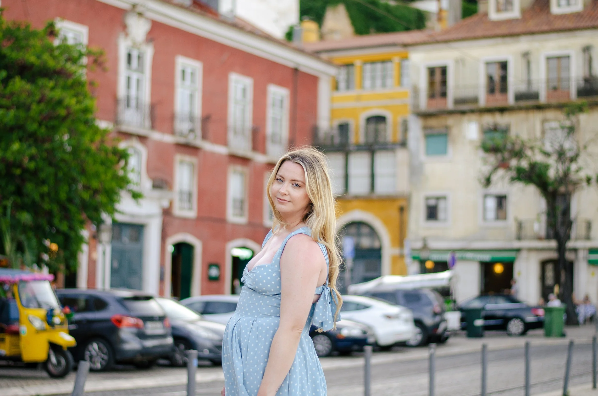 Teenage girl with blonde hair in a blue polka dot dress standing outdoors on a city street with colorful buildings and parked cars in the background.