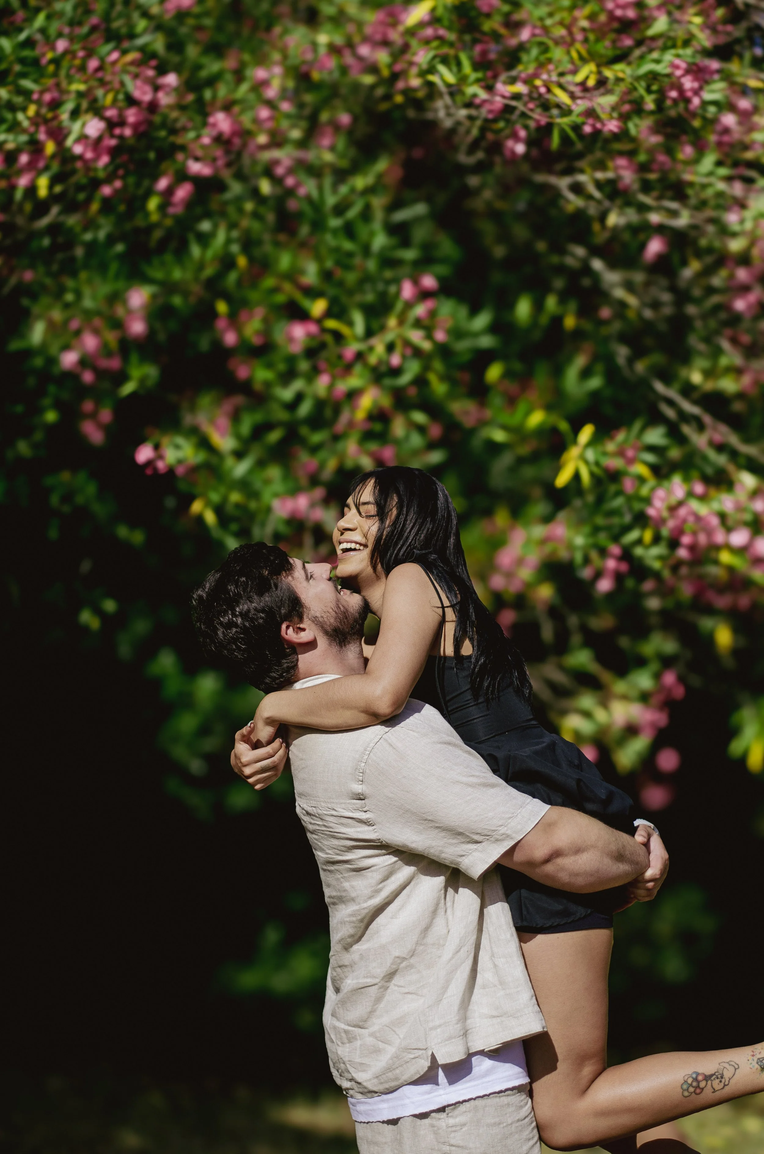 A man lifts a woman into the air while she laughs, with blooming pink flowers in the background.