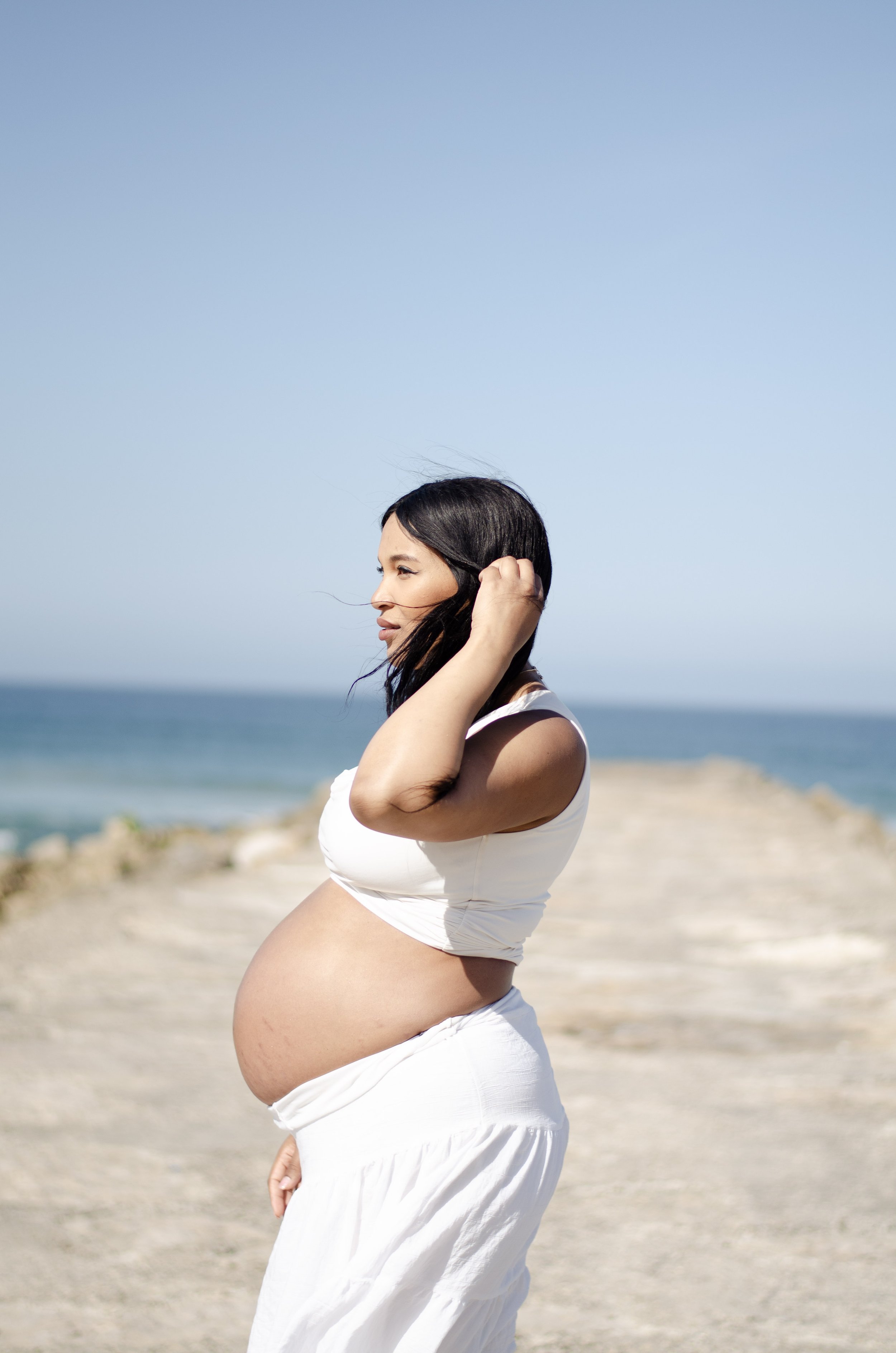 Pregnant woman wearing a white dress at the beach, with her hand touching her hair and standing in front of the ocean and a clear blue sky.