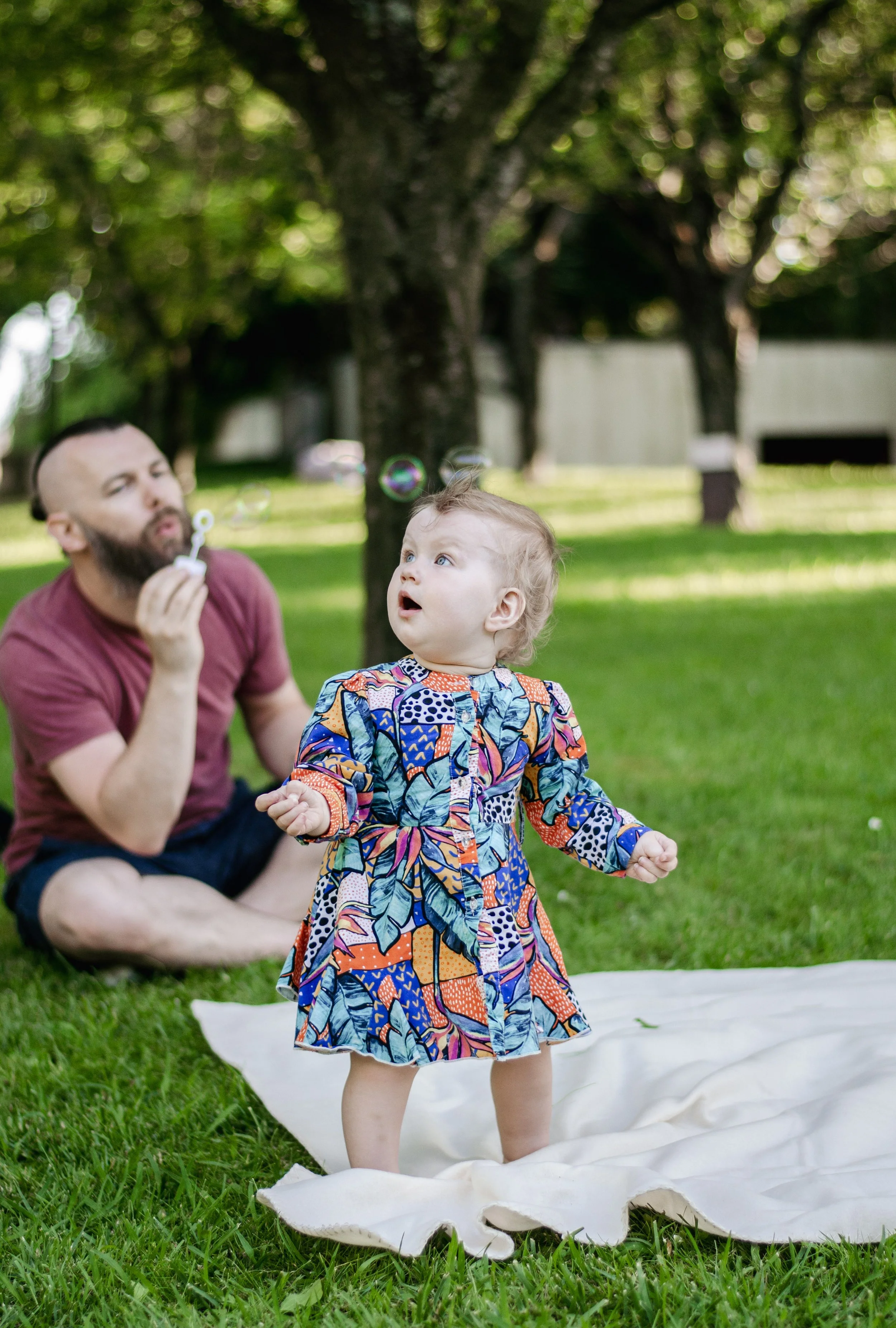 A young girl in a colorful patterned dress is standing on grass outdoors, reaching out with her hands, with an adult man in the background blowing bubbles.