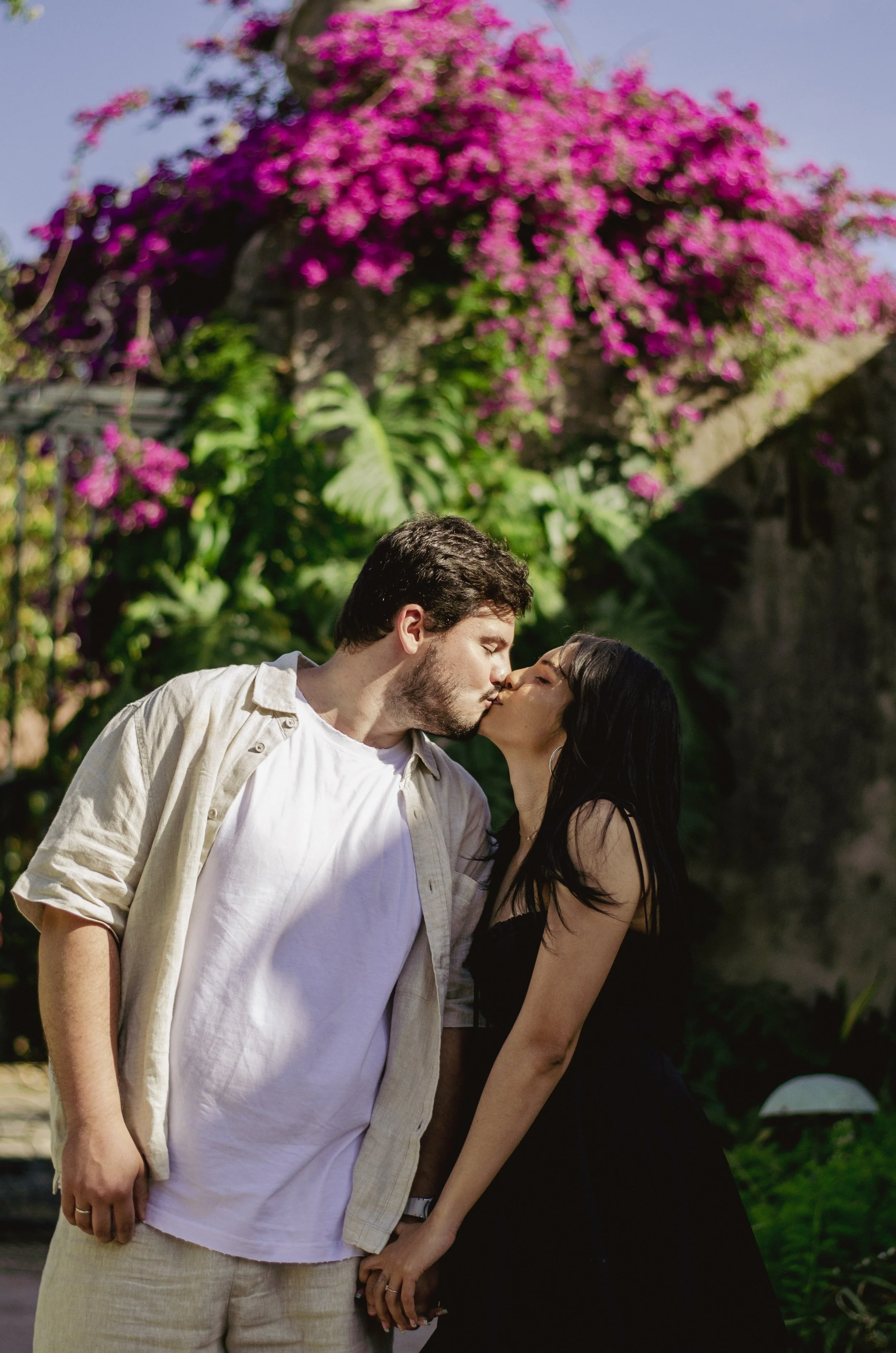 A man and woman kiss outdoors on a sunny day, with pink flowers and lush green plants in the background.
