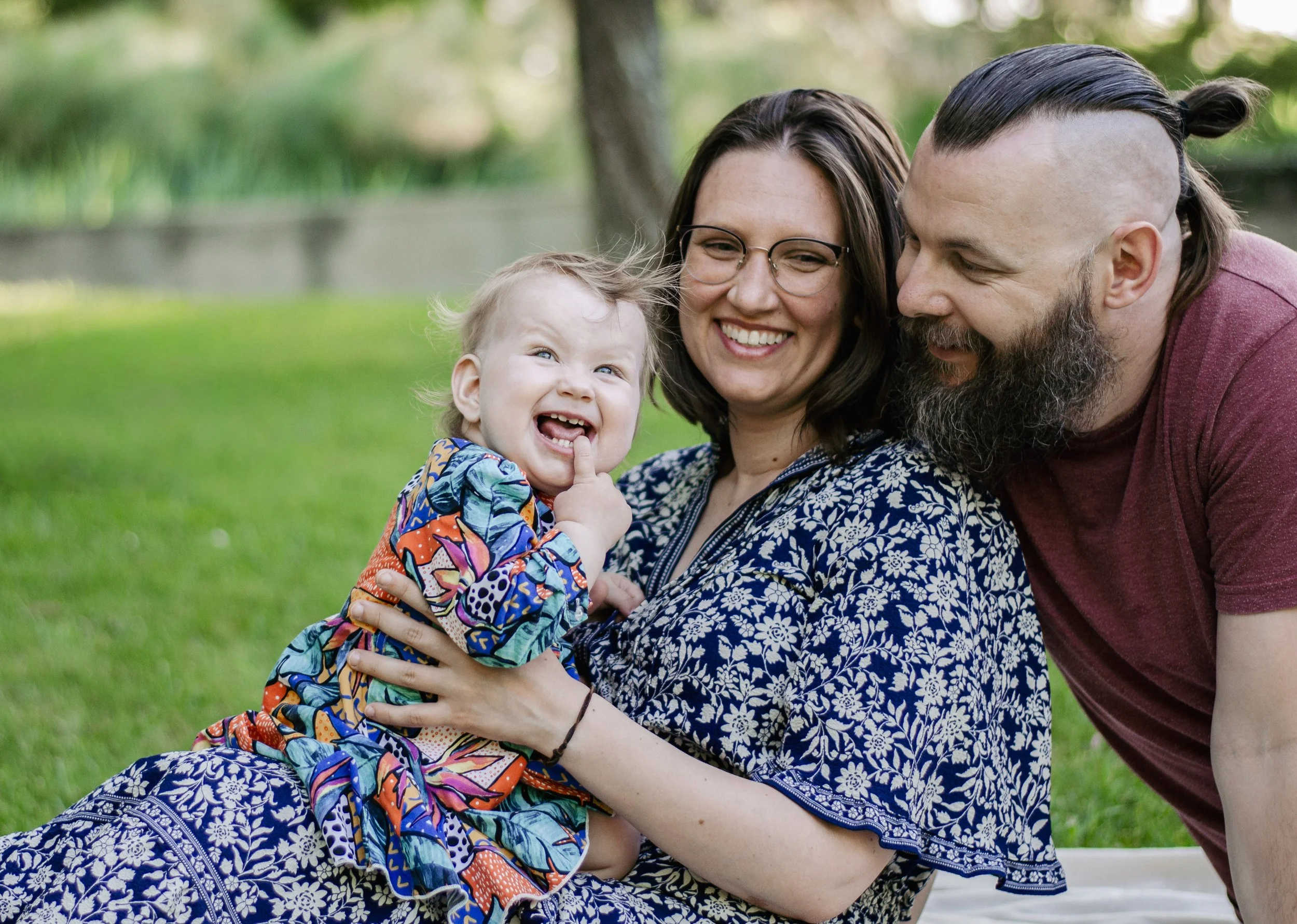 A family of three enjoying and playing together outdoors on a grassy area during daytime. The child is sitting on a woman's lap, smiling, with their finger in their mouth. The woman and man are close and smiling, with the woman holding the child and 
