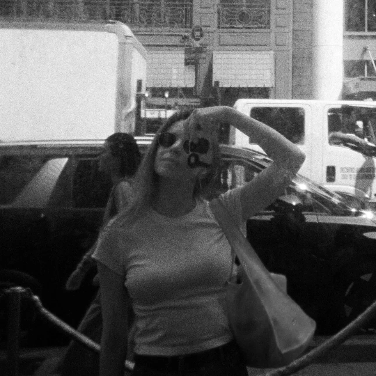 A woman taking a selfie in a mirror, wearing sunglasses and a light-colored shirt, with cars parked behind her and a cityscape in the background.