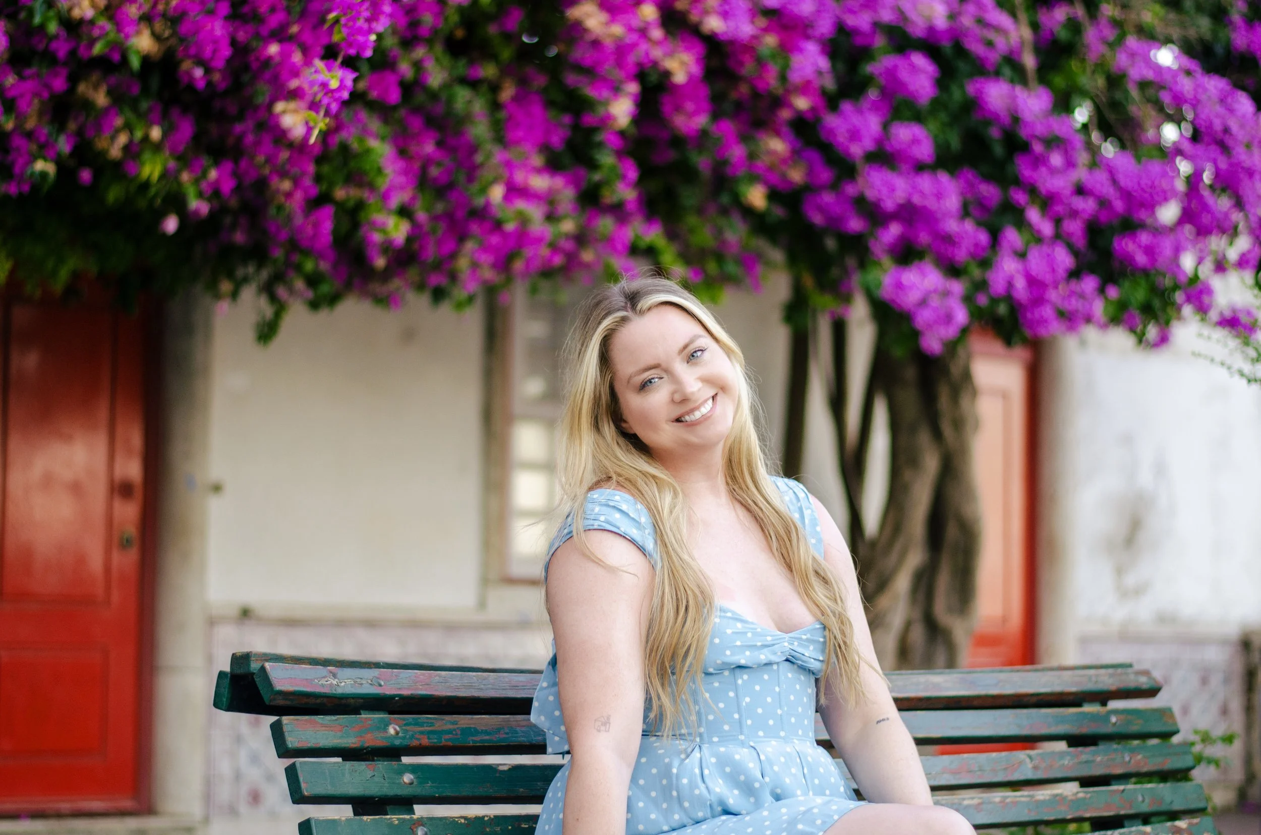 A young woman with long blonde hair and blue eyes sitting on a green park bench, smiling, in front of a tree with bright purple flowers, with a red door in the background.