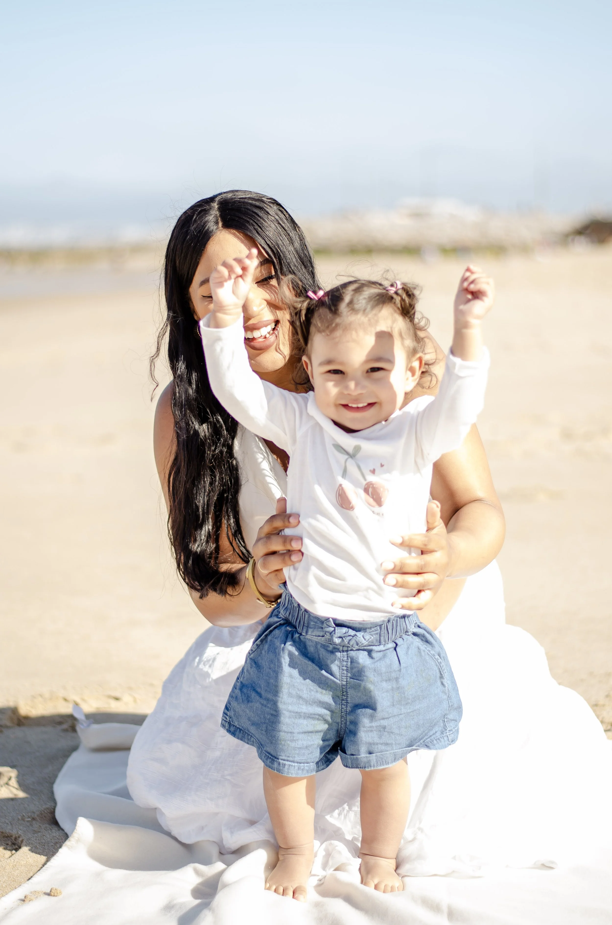 A woman with long black hair and a girl with curly hair in pigtails at the beach, smiling and raising their hands, standing on a white blanket on sand with a clear sky and blurred boats in the background.