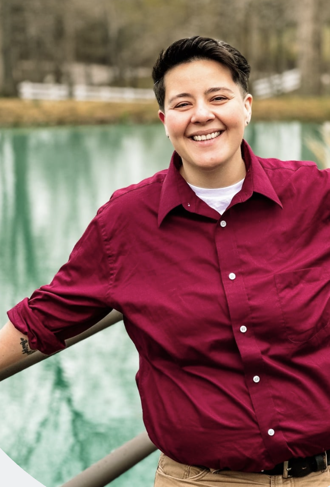 Smiling woman standing outdoors near a railing over a body of water, wearing a maroon button-up shirt, with trees and a white fence in the background.