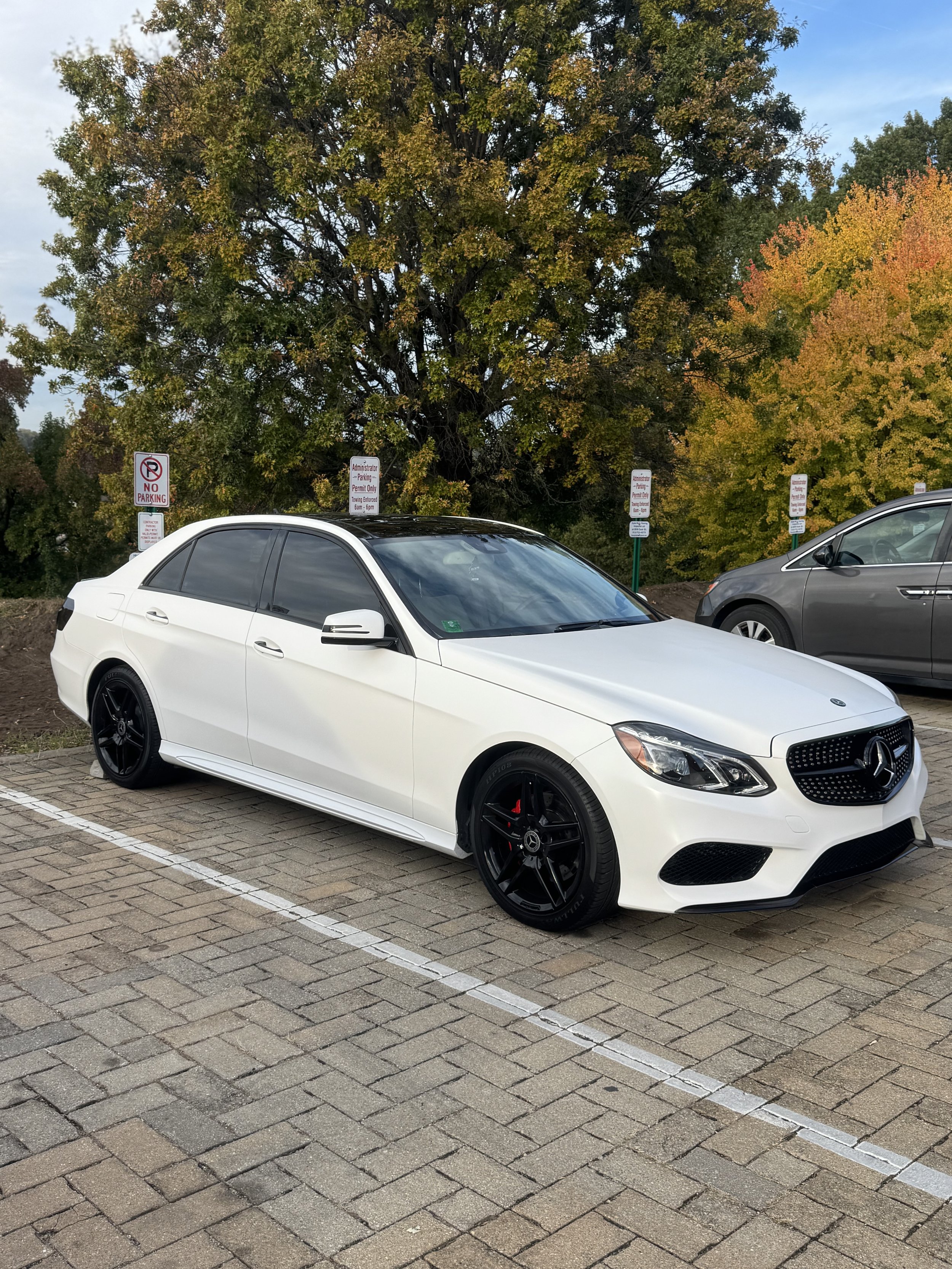 A white Mercedes-Benz parked in a parking lot with trees showing green and fall foliage in the background.
