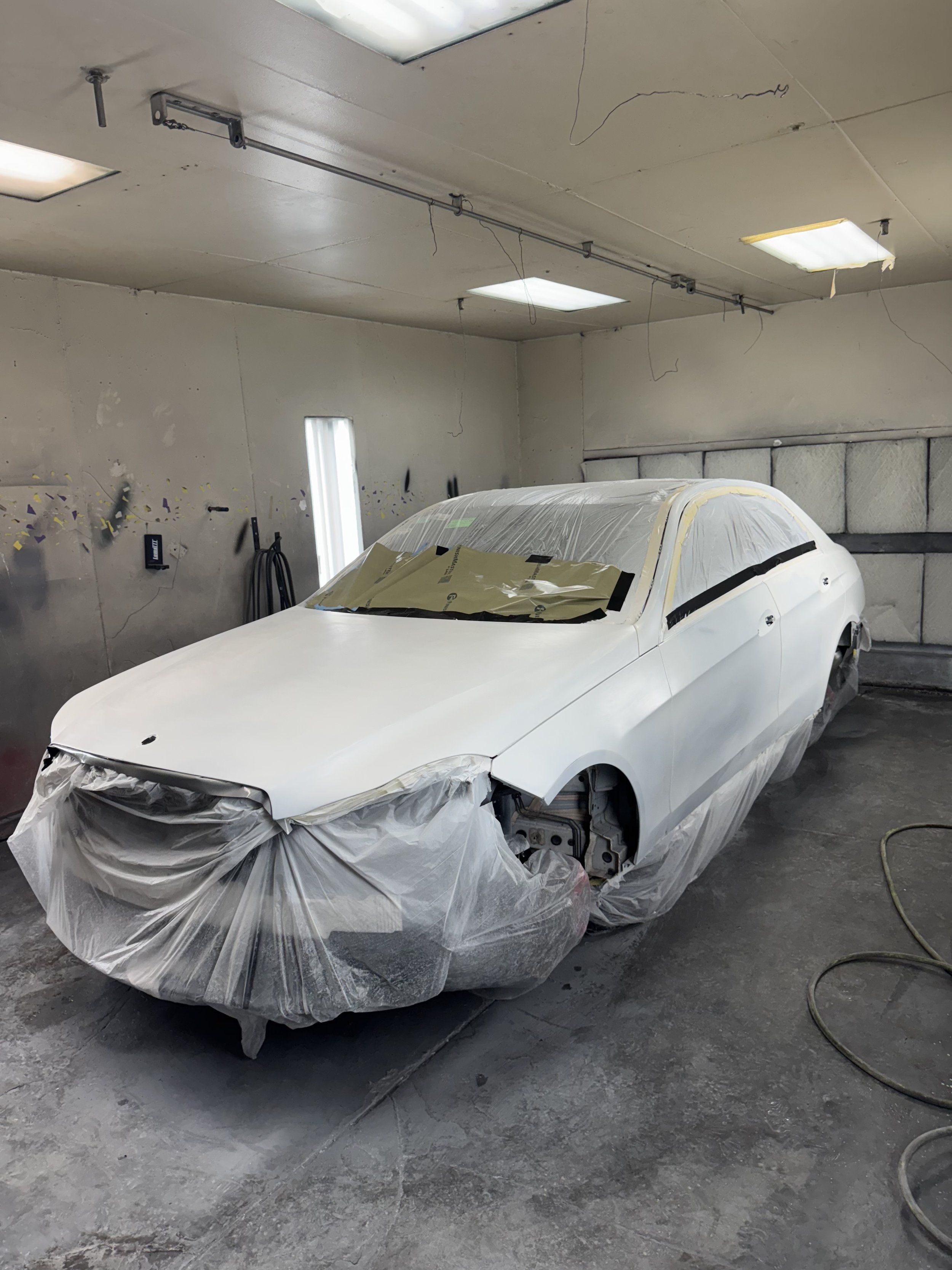 A white car in a workshop, covered in plastic sheeting for painting or repair, with parts of the front bumper removed. The indoor space has unfinished walls and ceiling with visible cracks and lighting fixtures.