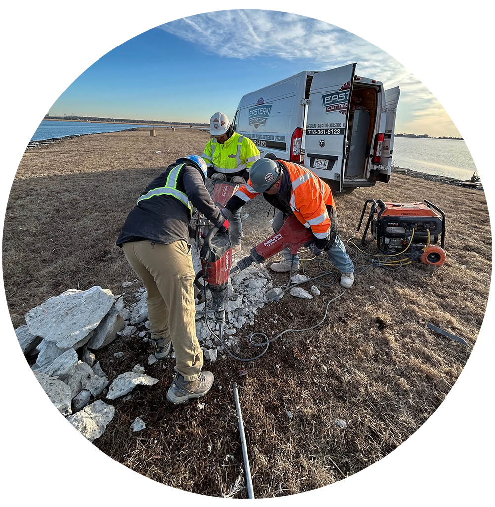 Eastern contractors using a jackhammer to break rocks on a grassy area near a body of water with an Eastern van in the background.