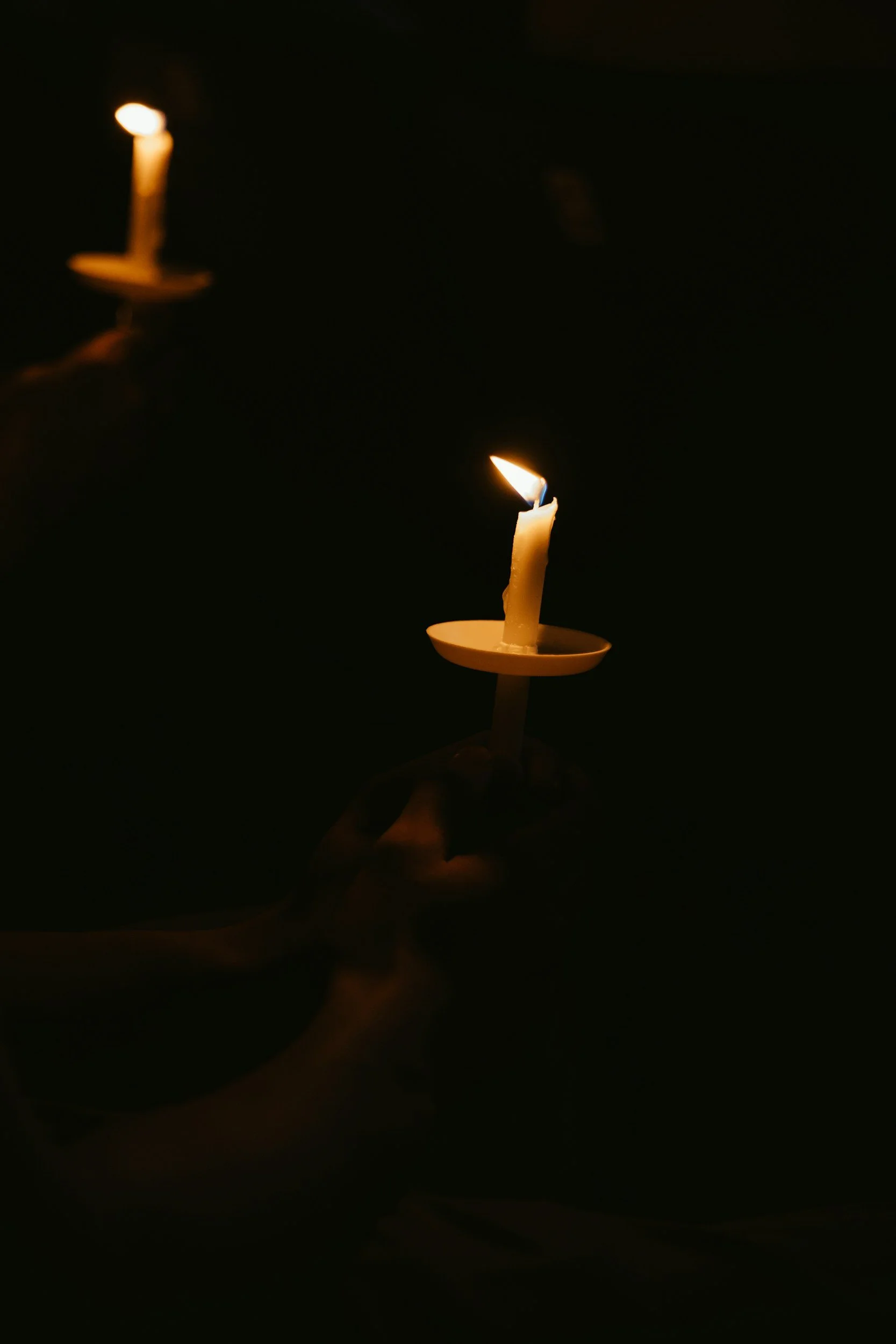 Two lit candles in dark surroundings, one being held by a hand.