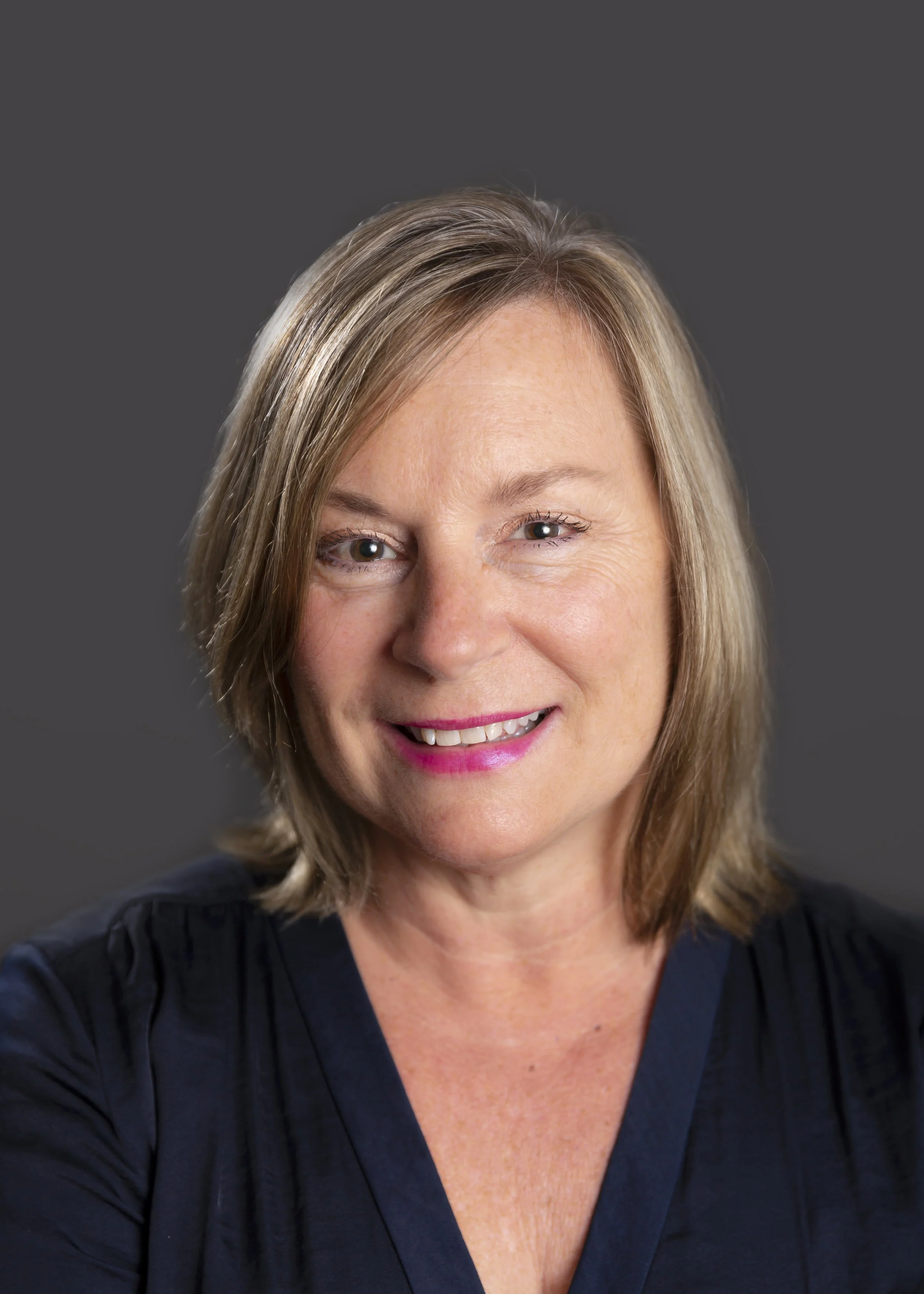 Close-up of a woman with shoulder-length blonde hair wearing a dark navy blue top, smiling, with a gray background.