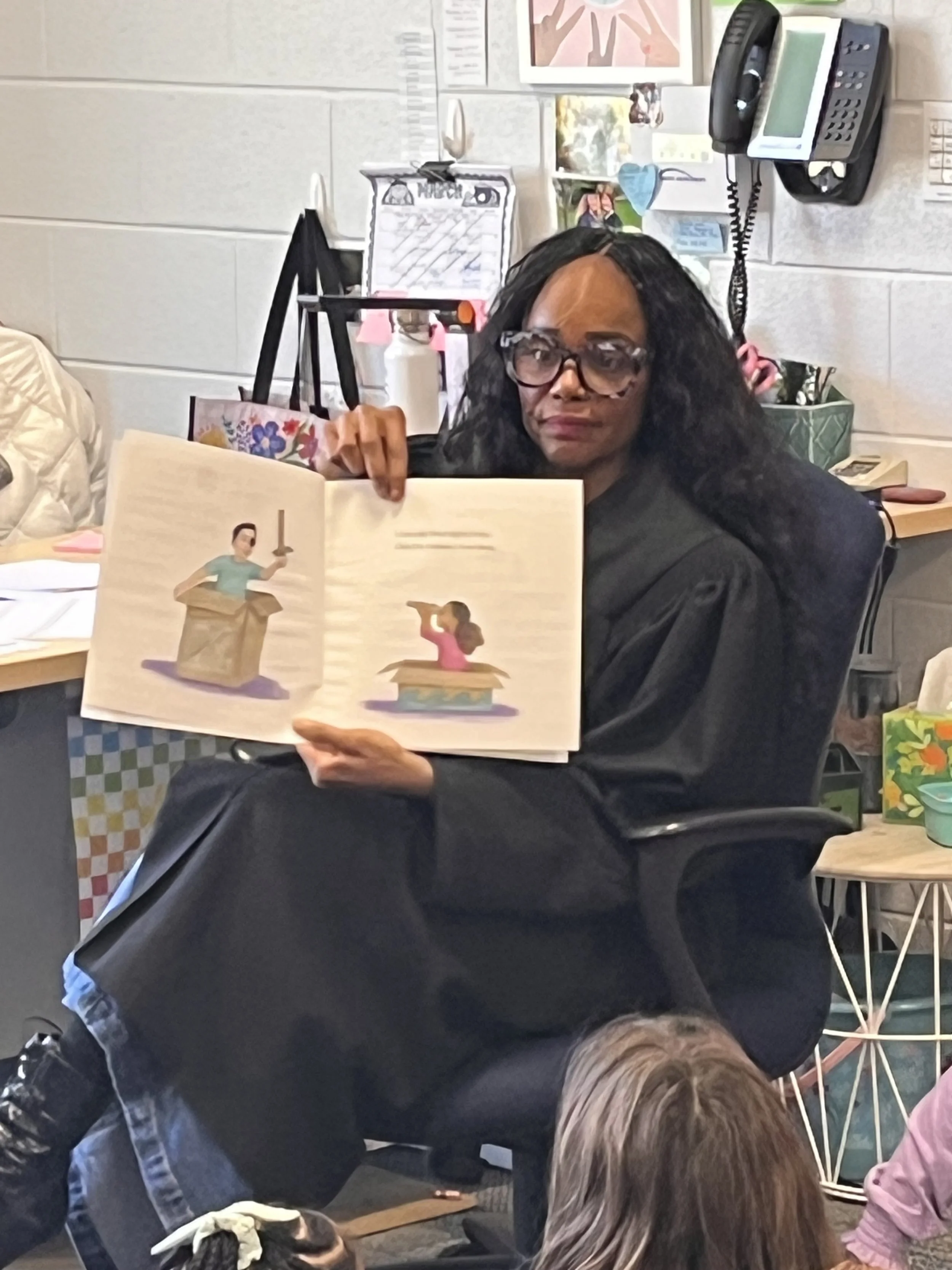 A woman with curly hair and glasses holding open a children's picture book with illustrations of a woman holding a sword and a girl standing on a book. She is sitting in a room with various office and art supplies, a phone, and framed pictures on the wall behind her.