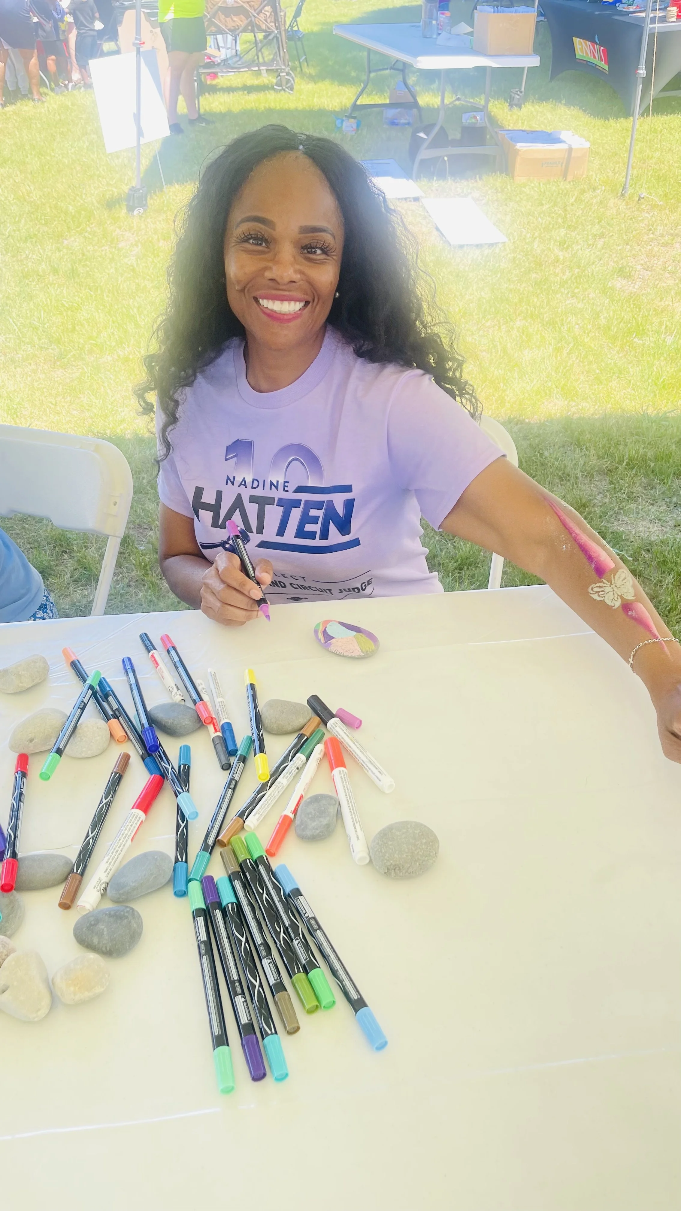 Smiling woman at a table covered with colorful markers and stones, painting a rock with pink and purple paint; outdoor event with tents and people in the background.