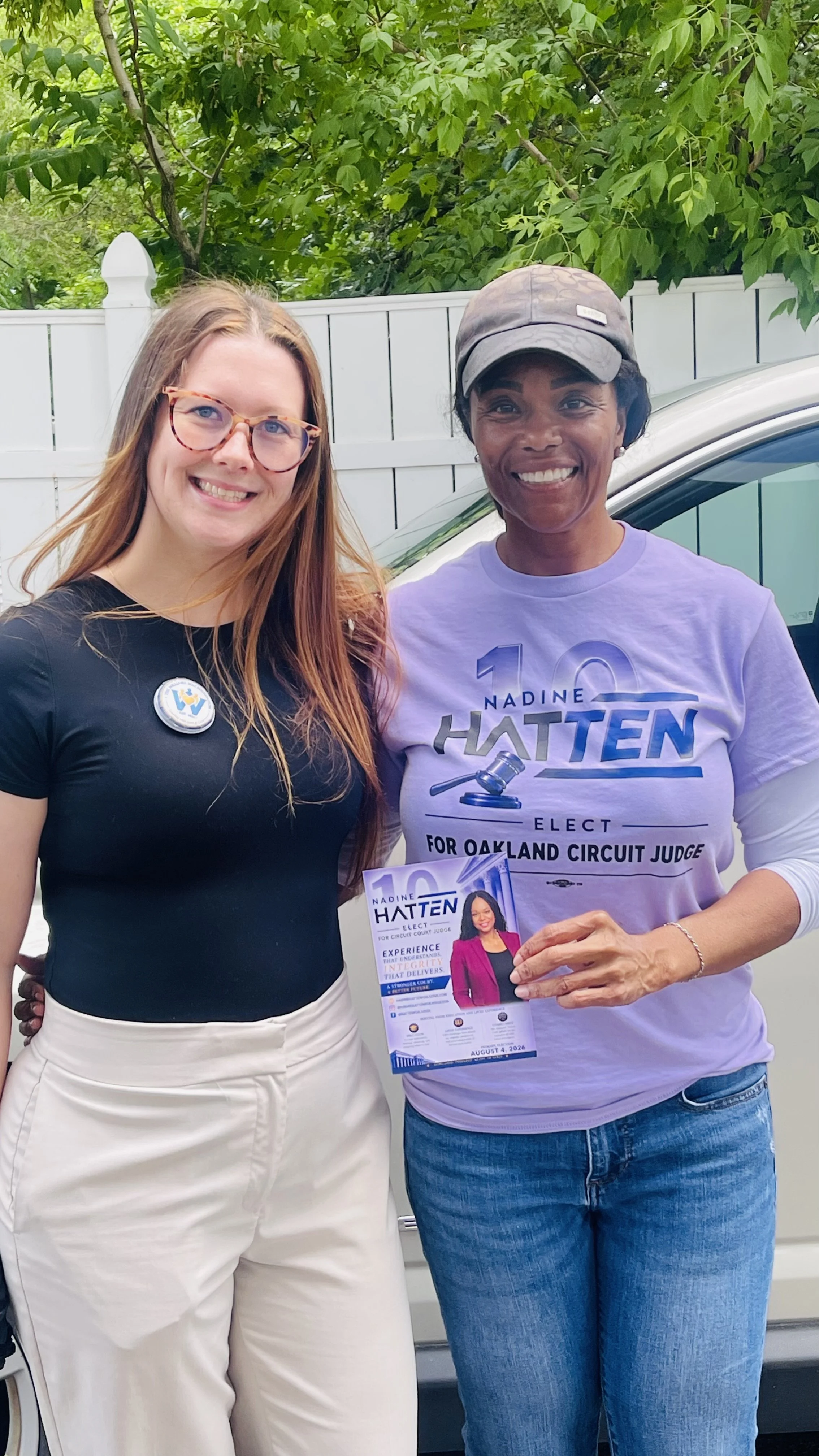 Two women standing outdoors in front of a white fence and green trees, smiling and holding campaign flyers. One woman has long red hair, glasses, a black top, and cream-colored pants. The other woman has short black hair, a gray cap, a lavender campaign shirt, and blue jeans.