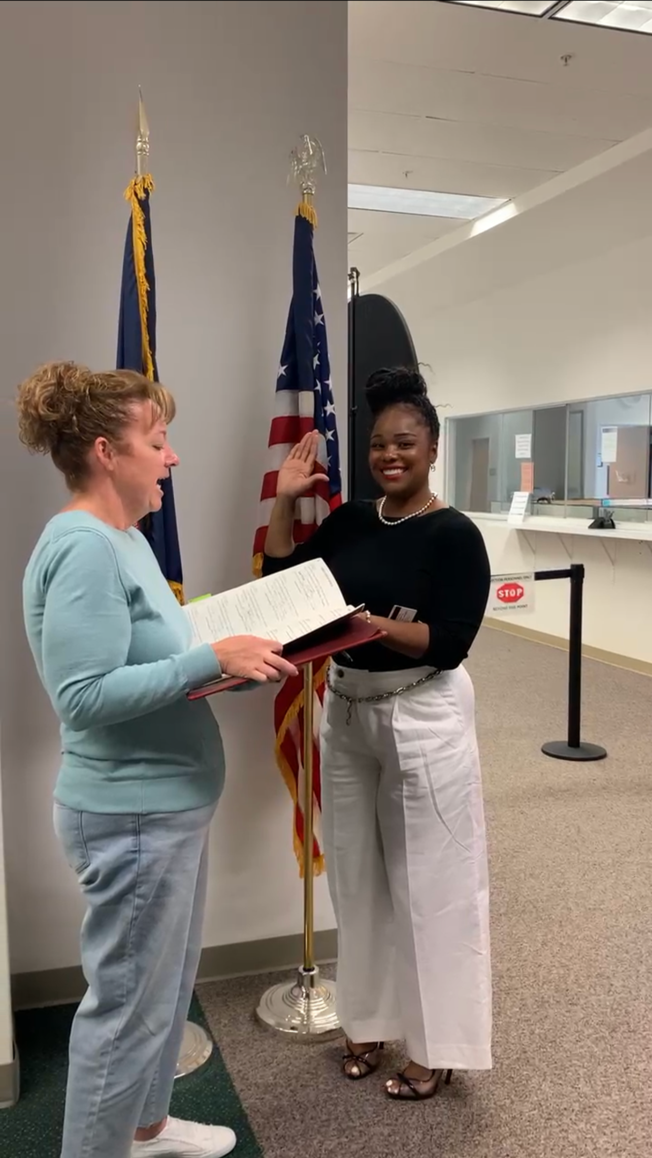 A woman taking an oath with her right hand raised, standing next to another woman reading from a document, near U.S. flags in an indoor setting.
