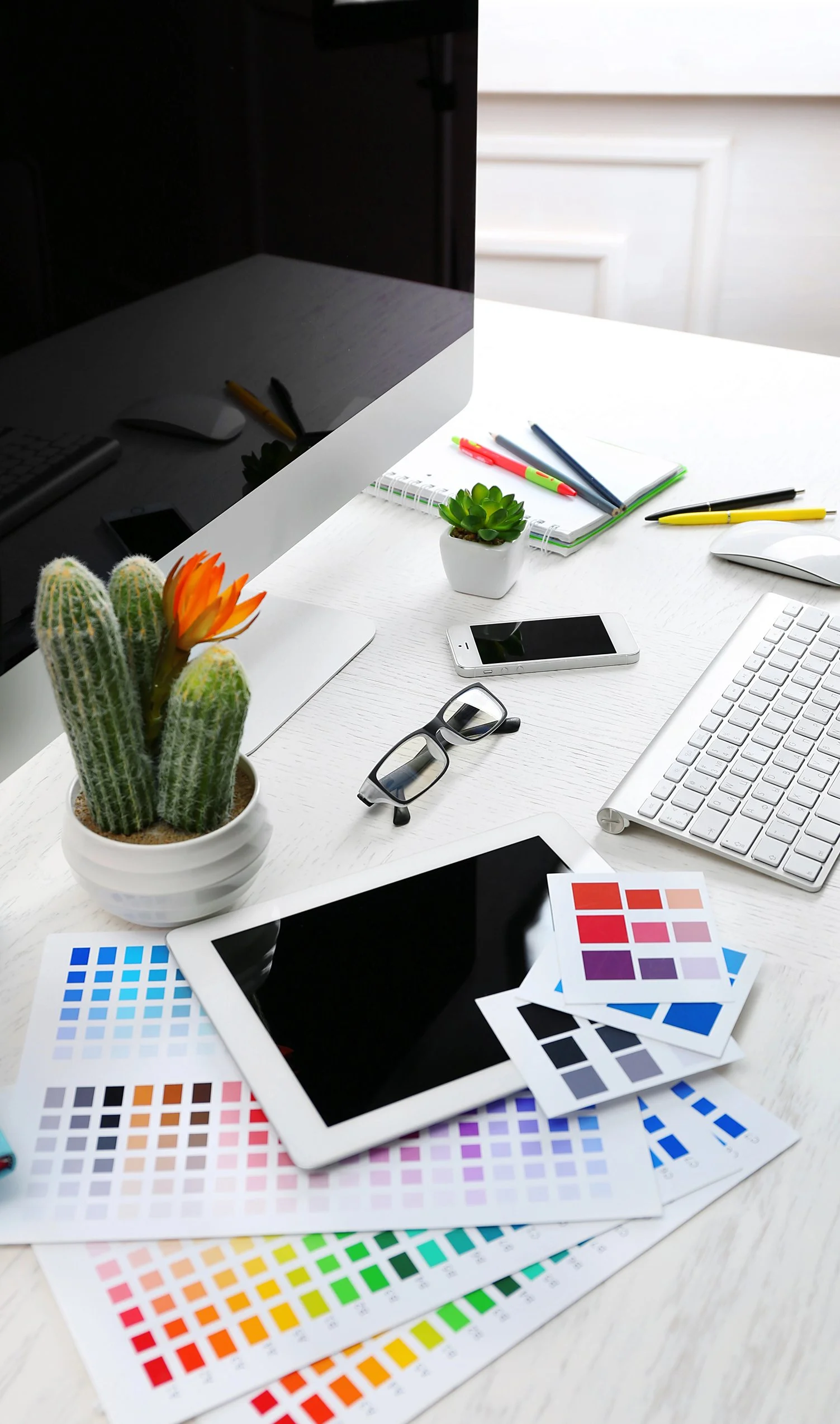 Desk with cactus plant, color swatches, tablet, glasses, smartphone, notepad, pens, keyboard, mouse, and computer monitor