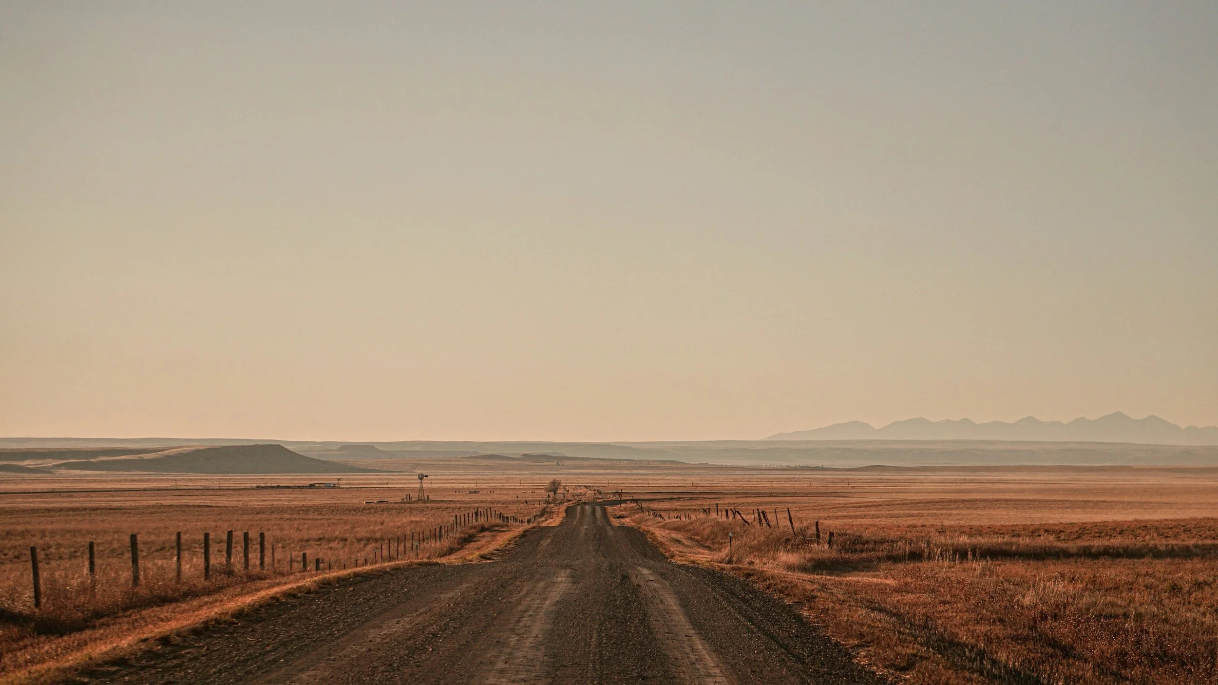A rural dirt road stretches into the distance through open plains, with mountains visible on the horizon and a fence on both sides of the road.