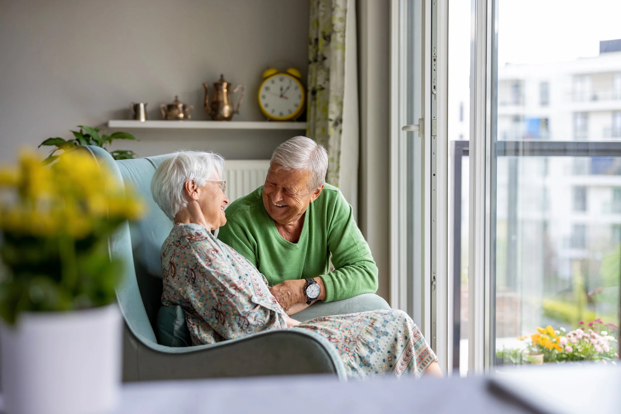 An elderly woman and elderly man sitting together on a sofa in a bright living room, sharing a happy moment and smiling at each other. The woman is wearing glasses and a floral dress, while the man is wearing a green sweater and a watch on his wrist. There are flowers on a table in the foreground, and a window with a view of an apartment building behind them.