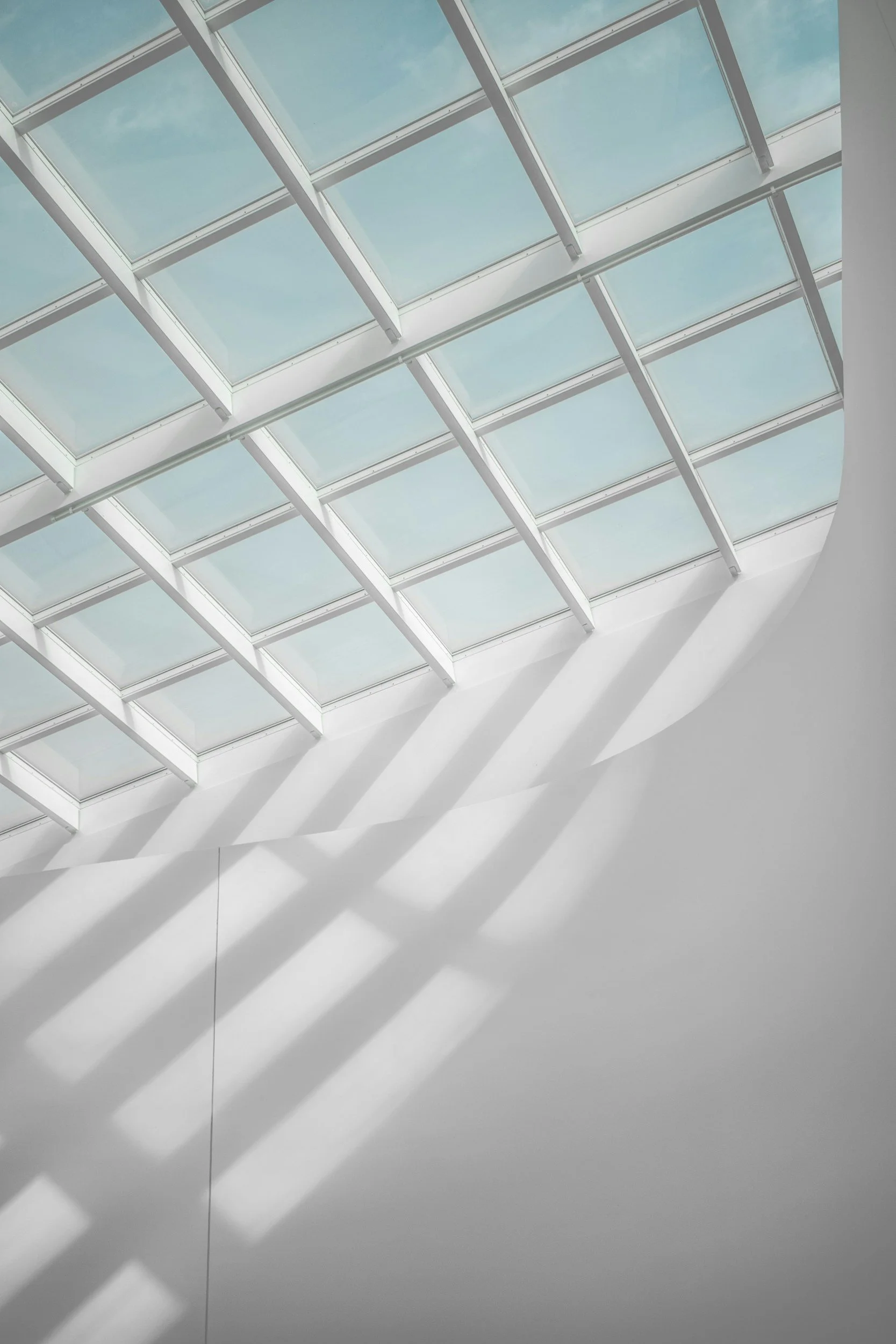 A ceiling with glass panels, showing the sky through them, and shadows of the panels cast on a white wall below.