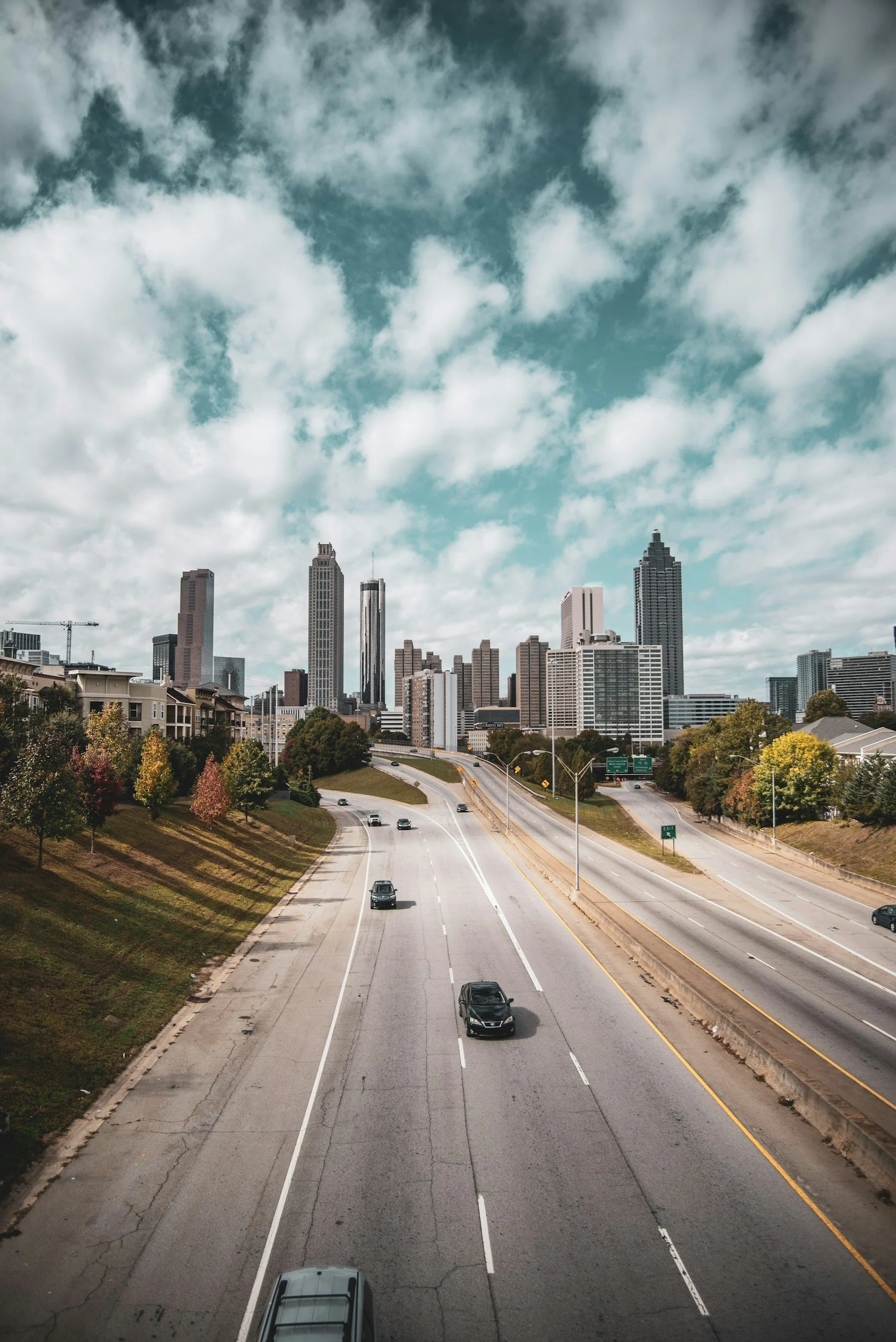 A city skyline viewed from a multi-lane highway with a few cars, with skyscrapers under a partly cloudy sky.