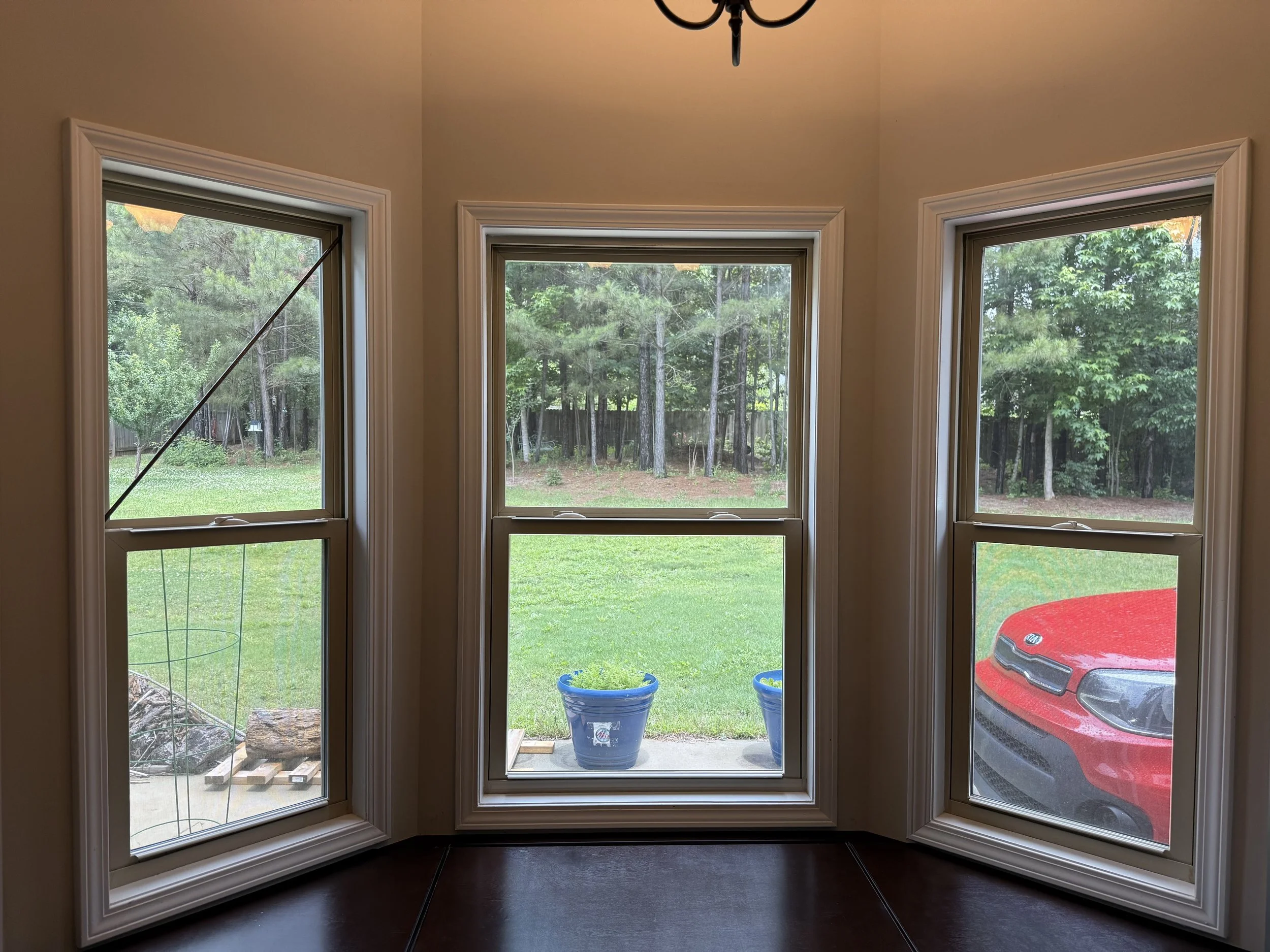 A view from inside a house showing three large windows overlooking a backyard with trees, a grassy lawn, and a red vehicle parked outside. The middle window has two blue planters on the porch, and the left window has a diagonal support rod.
