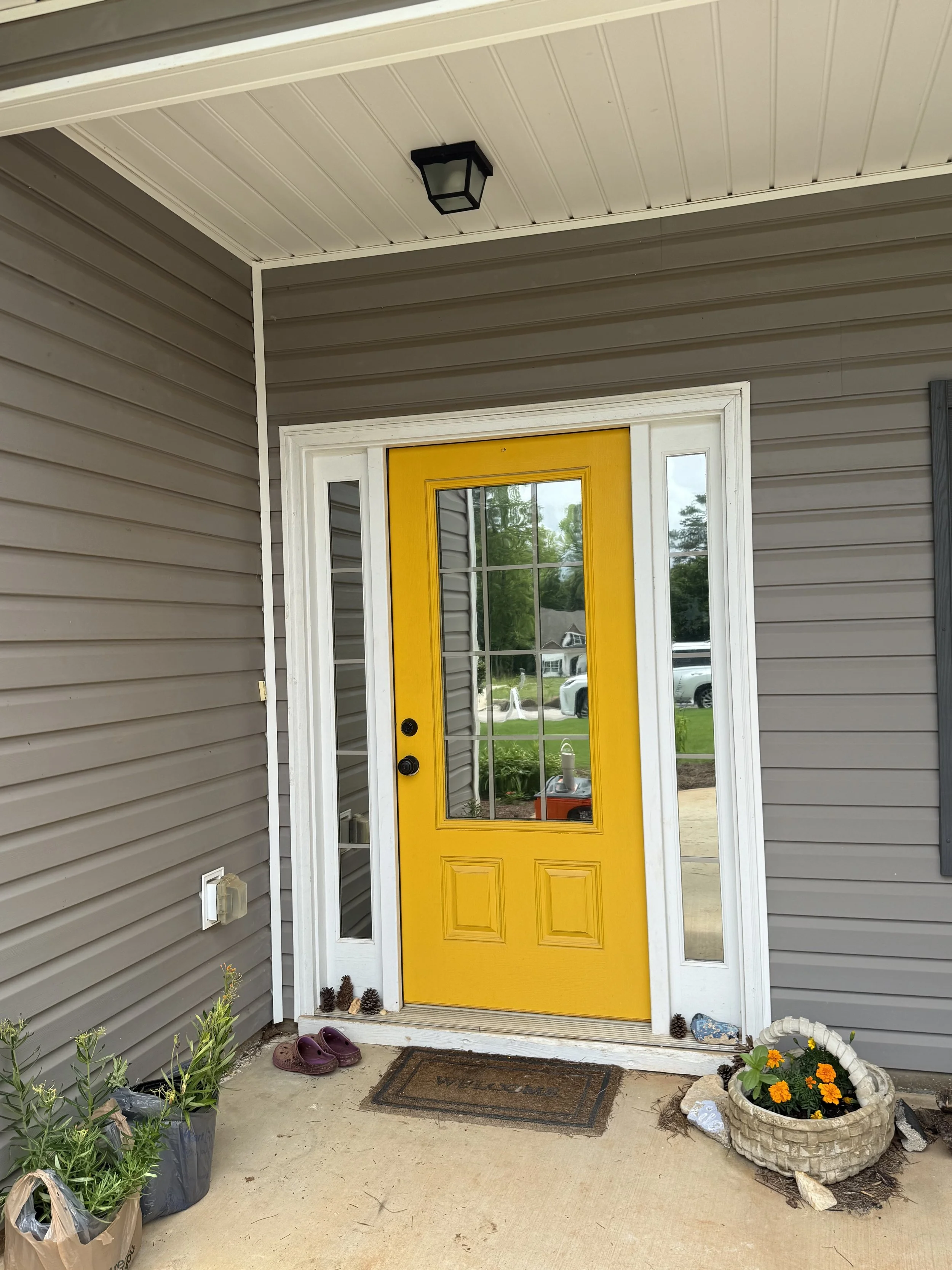 Front porch area with a yellow door and side windows, gray vinyl siding, purple slippers on the ground, and a doormat. There are plants in pots, pinecones, and a flower basket beside the door, with some decorative rocks and mulch on the soil.
