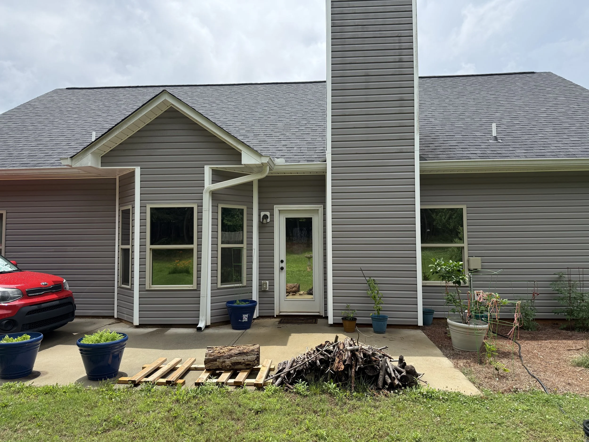 The back of a house with gray siding, a pitched roof, and multiple windows. There are potted plants and gardening supplies on the patio, with cloudy skies overhead.