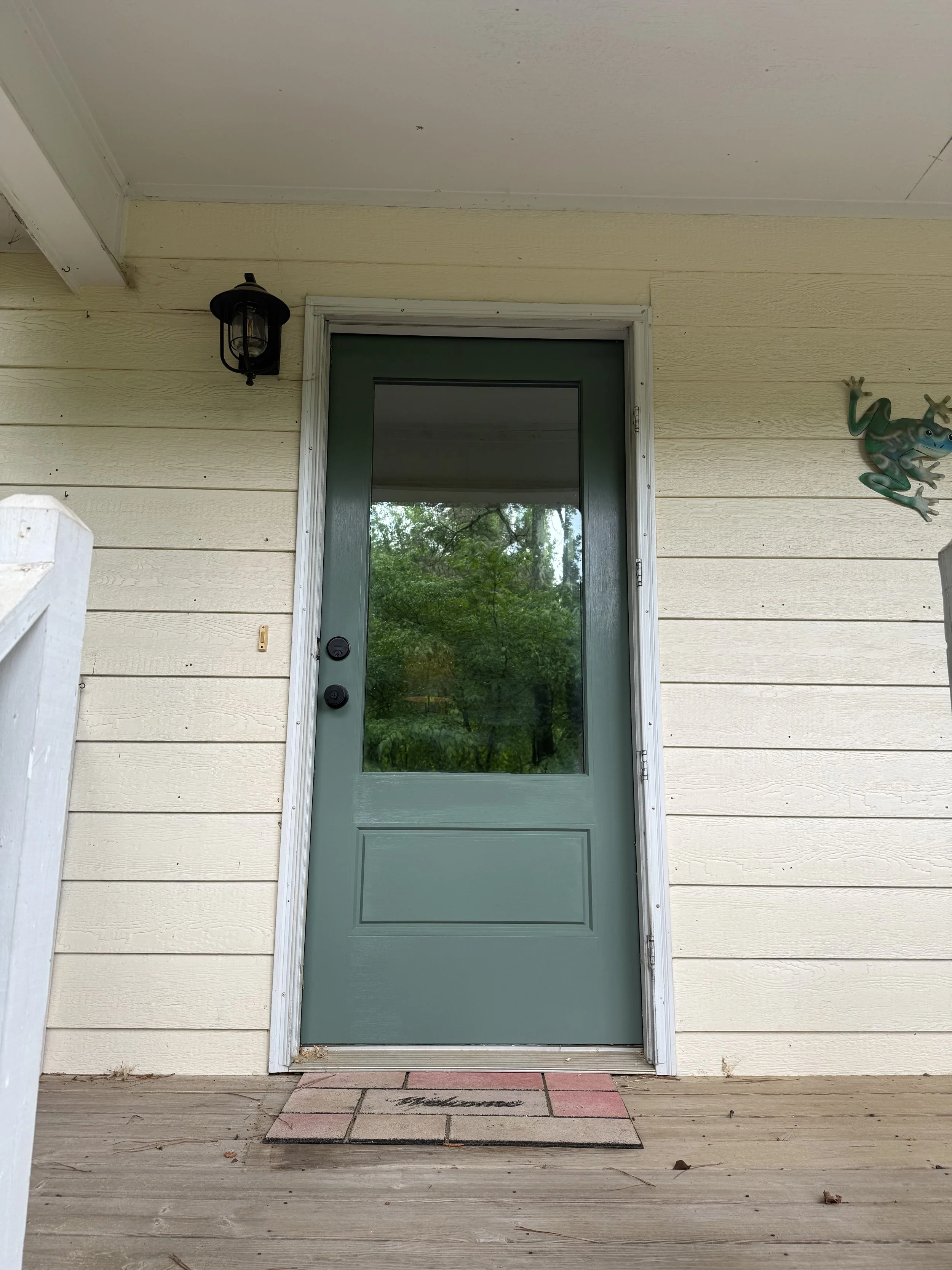Front porch with beige siding, green door with glass window, black outdoor lantern above the door, decorative frog wall sculpture to the right, and a welcome mat on the wooden porch floor.