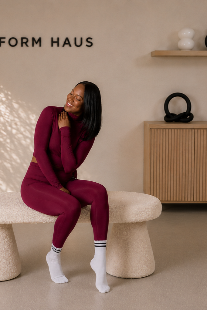 A woman in a matching maroon sportswear set sitting on an off-white textured bench with her eyes closed and a broad smile, in a modern interior with a beige wall, nearby a wooden cabinet with black decorative object, and a wall shelf with white vases.