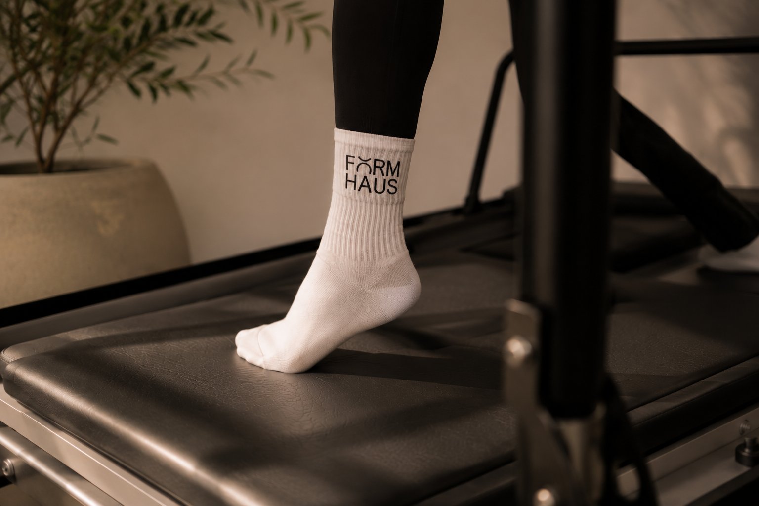 A person's foot and ankle wearing a white sock with the words 'FÖRM HAUS' on it, standing on a black padded rehabilitation or treadmill machine indoors.