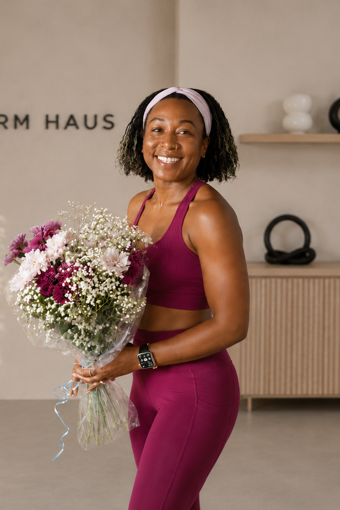 A woman in athletic wear holds a bouquet of pink and white flowers, smiling, inside a modern fitness studio.