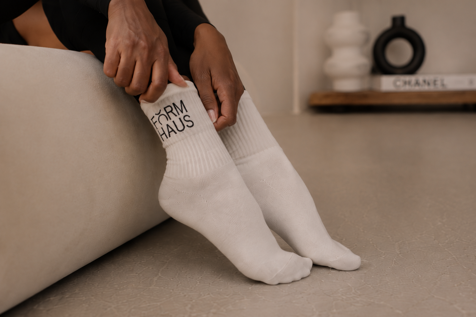 Person adjusting white socks with 'FORM HAUS' written on the cuff, sitting on a beige sofa in a modern room with a wooden shelf and decorative items.