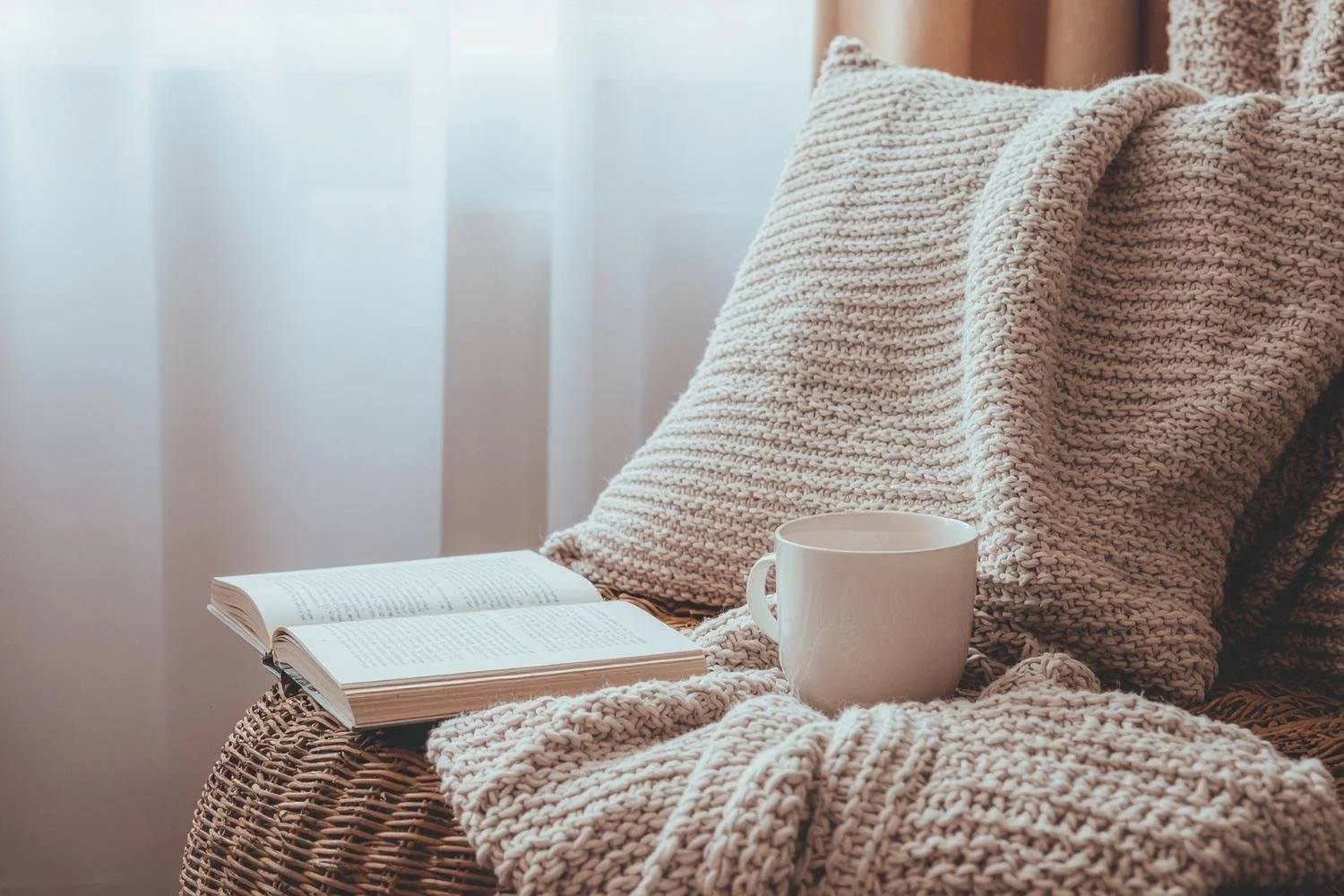 A cozy scene with a knitted blanket, an open book, and a white mug on a wicker chair near a window with sheer curtains.