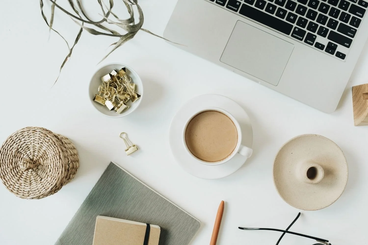 Top view of a white desk with a laptop, a cup of coffee, a notebook, a pen, and a pair of eyeglasses.