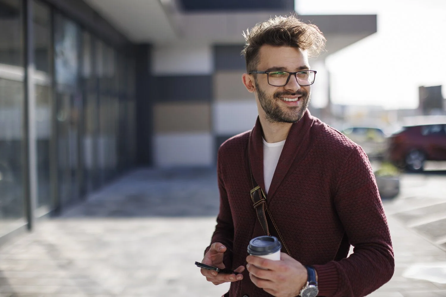 Young man with glasses, beard, and curly hair smiling, holding a coffee cup and smartphone outside on a sunny day.