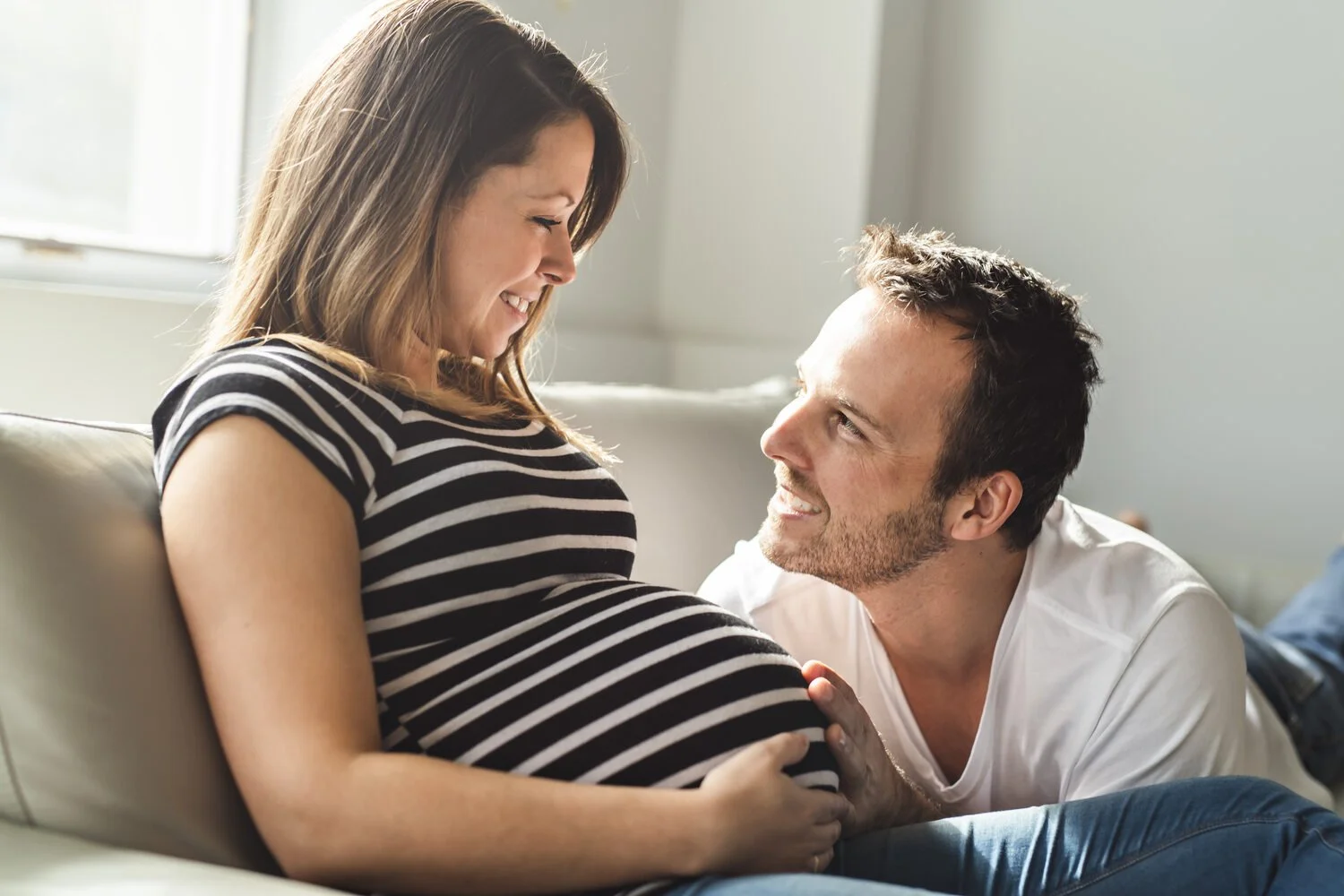 A pregnant woman smiling at her partner while sitting on a couch, with her hand on her belly and her partner's hand resting on her tummy.