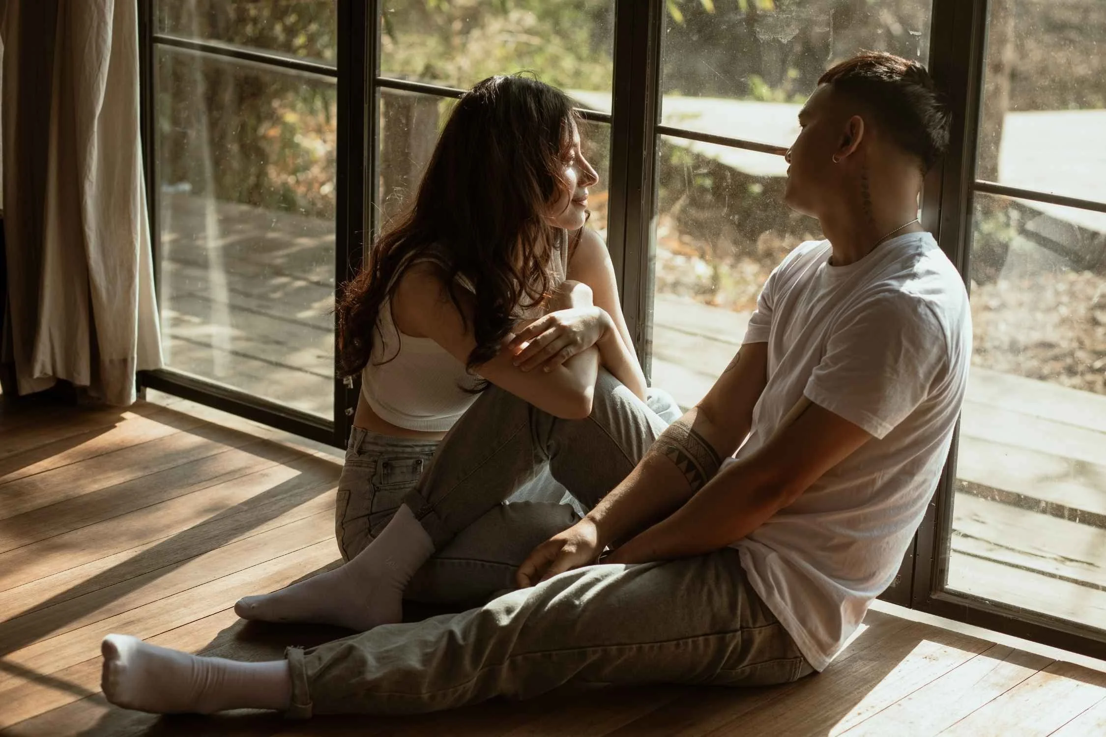 A couple sitting on the wooden floor by a large window with natural light, the woman smiling and the man looking at her.