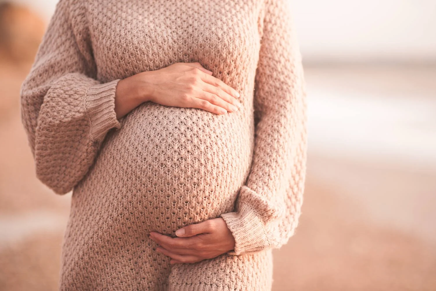 A pregnant woman wearing a knit sweater, cradling her baby bump with one hand on her stomach and the other on her chest.