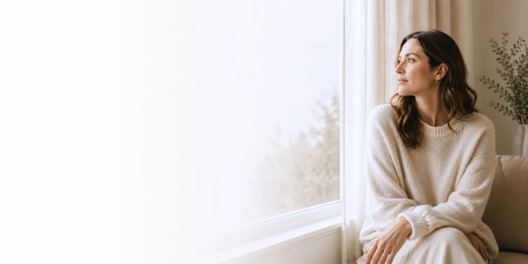 A woman sitting by a window with a view outside, wearing a cream-colored sweater, looking thoughtfully out the window.