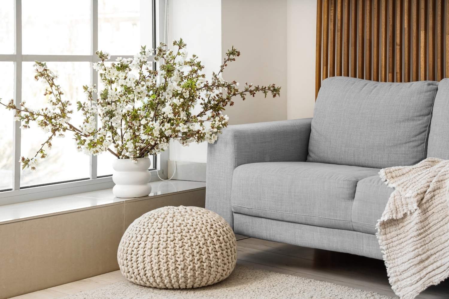 A cozy living room corner with a light gray sofa, beige knitted pouf, a white vase with white flowering branches on the windowsill.