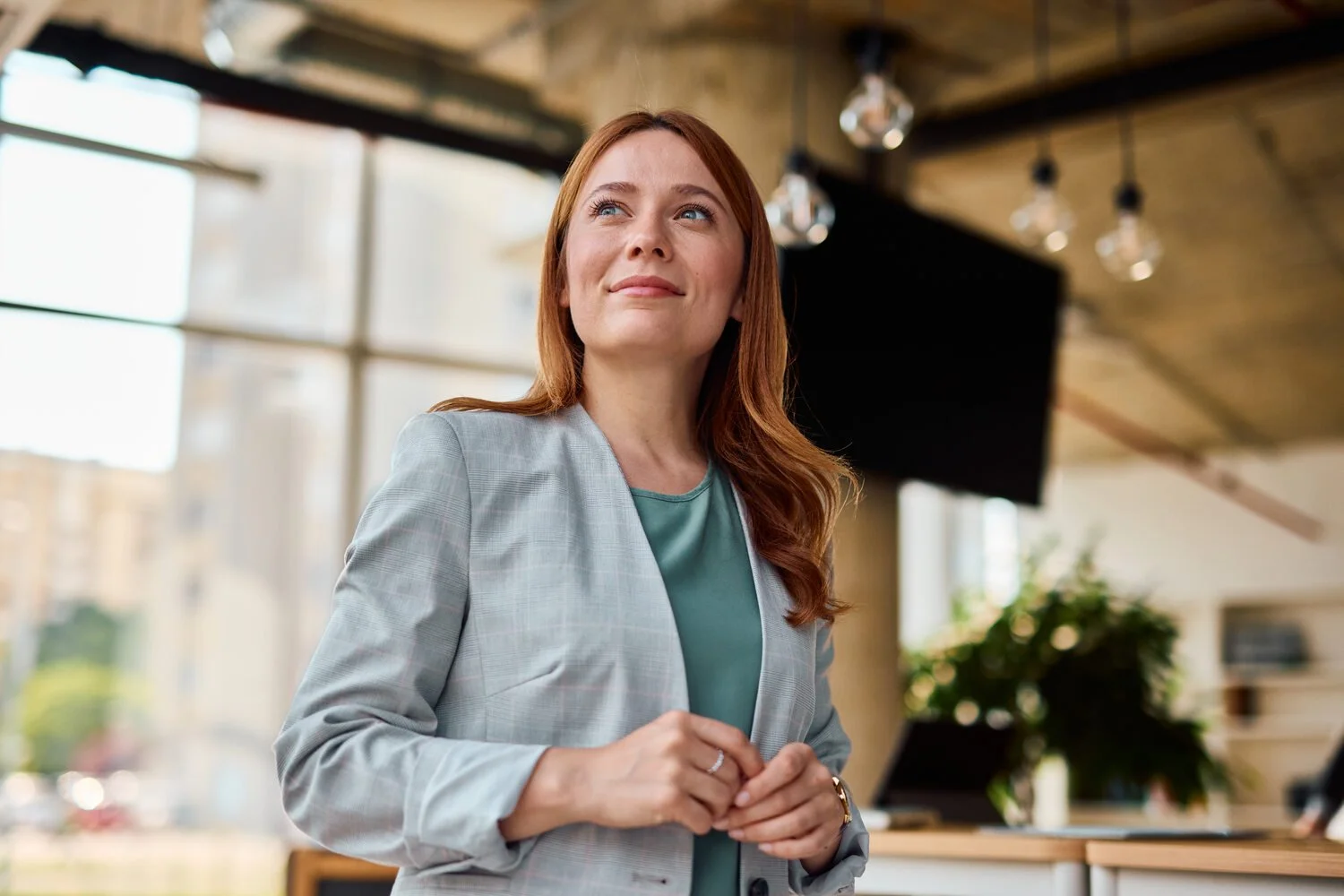 A woman with red hair wearing a light gray blazer and a teal top, smiling and standing in a modern office.