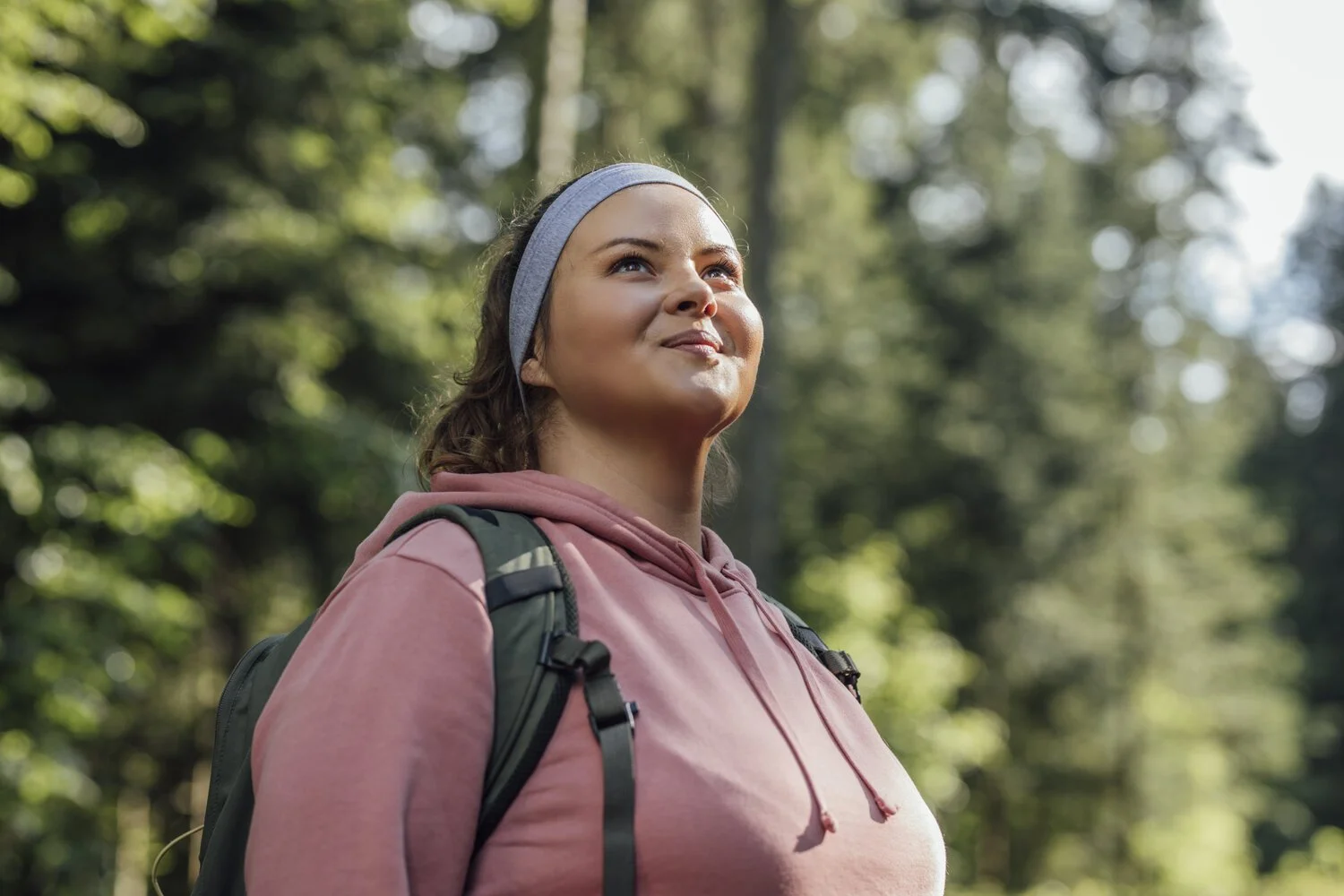 A woman with a backpack, wearing a pink hoodie and a headband, standing outdoors in a forest, looking up with a smile.