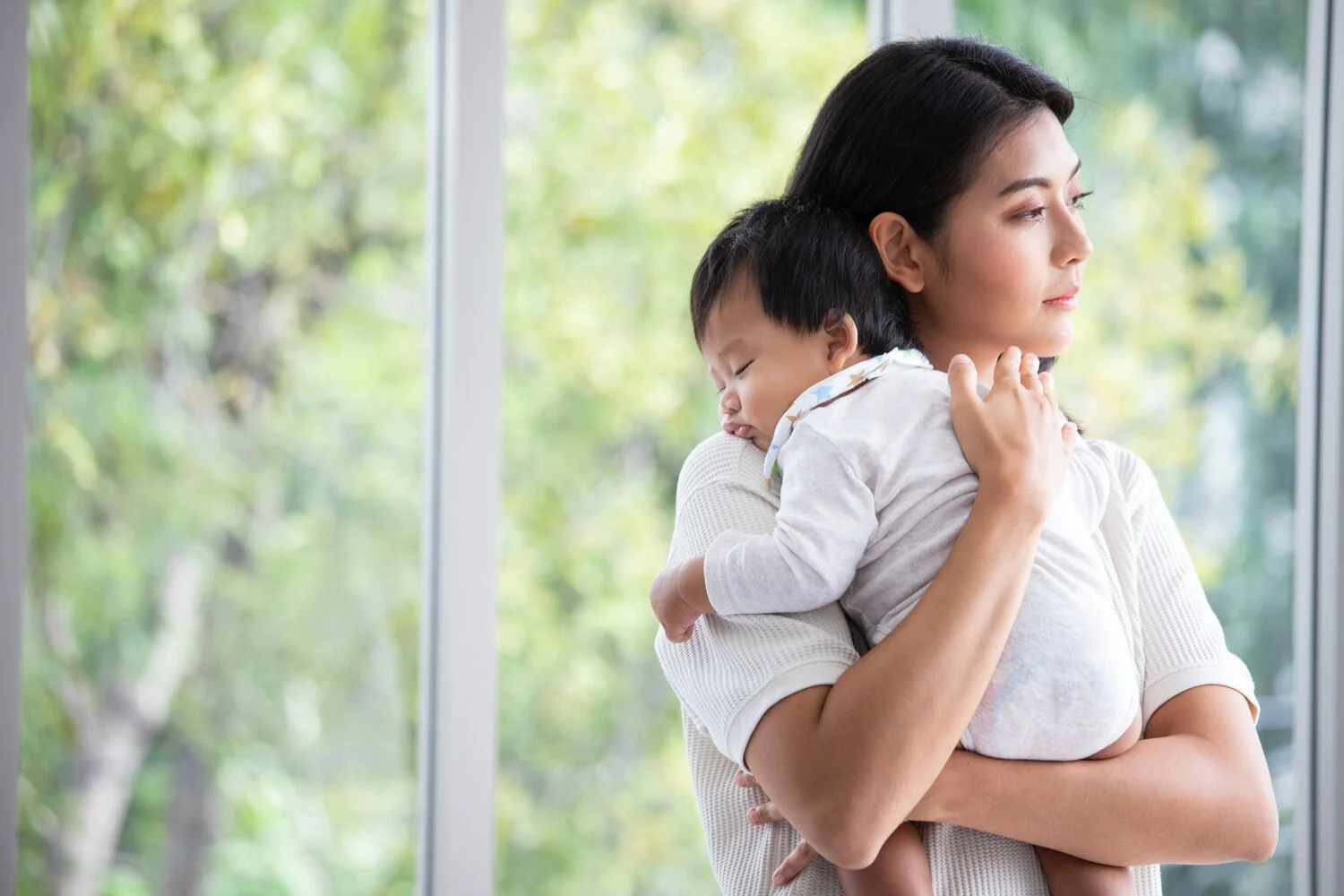 A woman holding a young child in her arms, standing by a large window outside view of green trees.