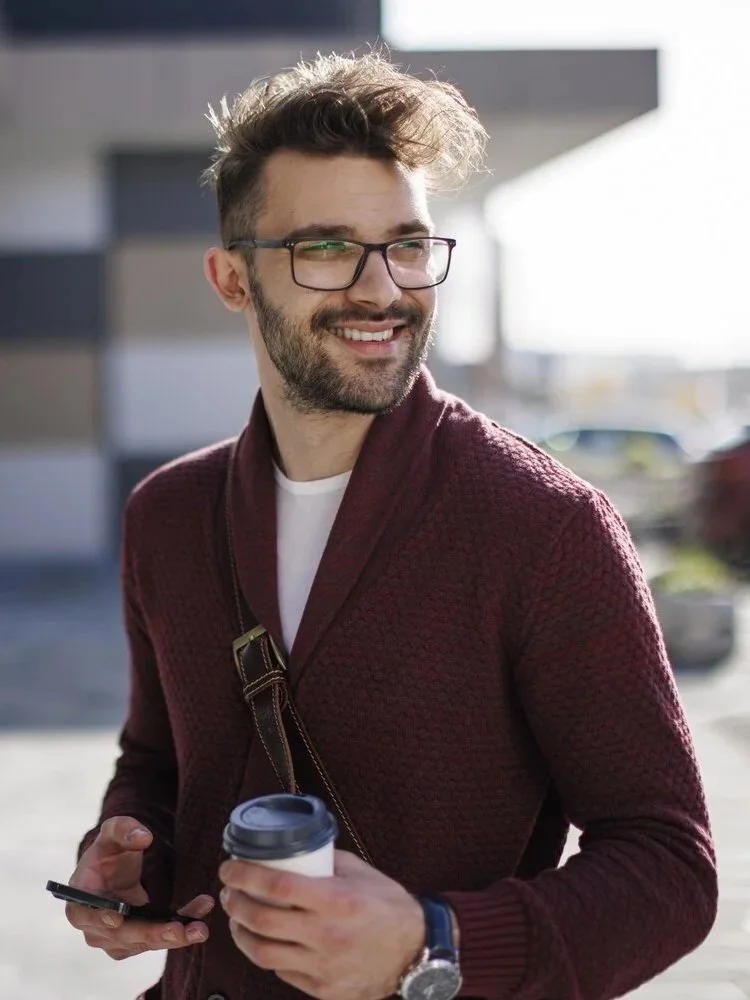 Young man with glasses, beard, and curly hair smiling, holding a coffee cup and smartphone outside on a sunny day.