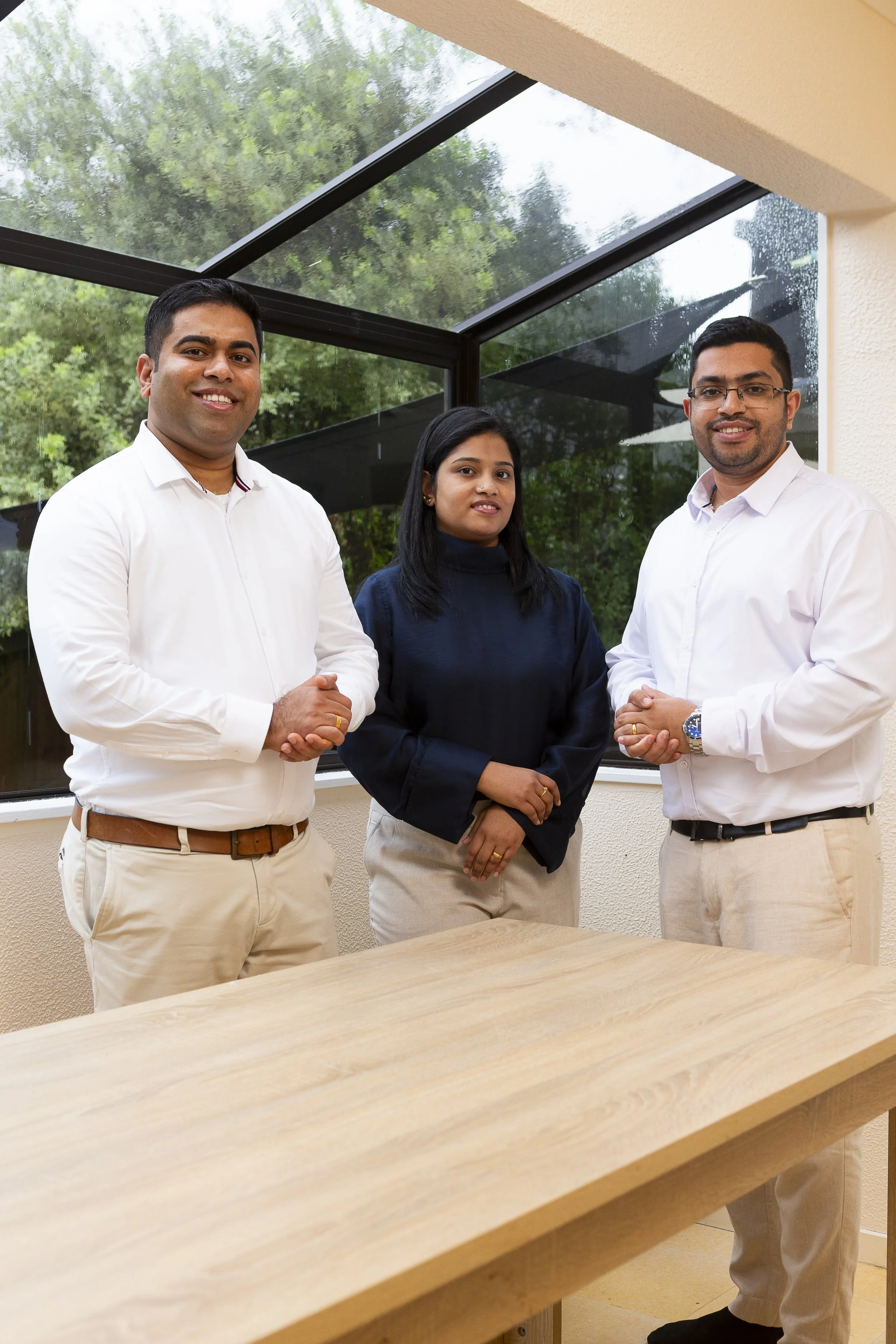 Three professionals posing for a photo indoors by a large window with greenery outside. Two men are standing on either side of a woman, all dressed in business casual attire, smiling.