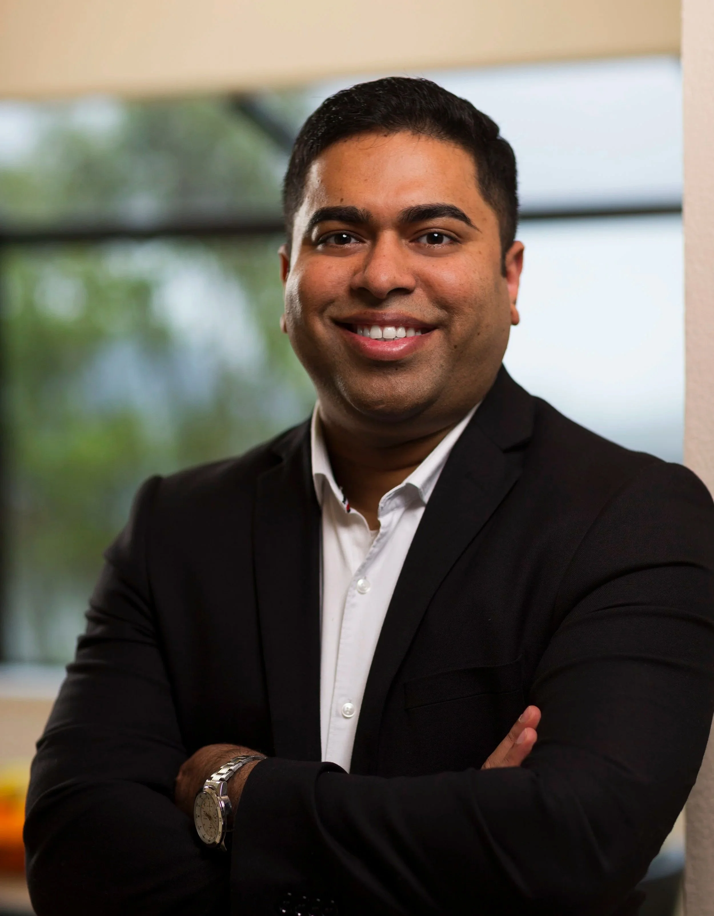 Portrait of a smiling man wearing a black suit, white shirt, and a wristwatch, with crossed arms, indoor background with large windows and blurred scenery. Mortgage advisor in Auckland