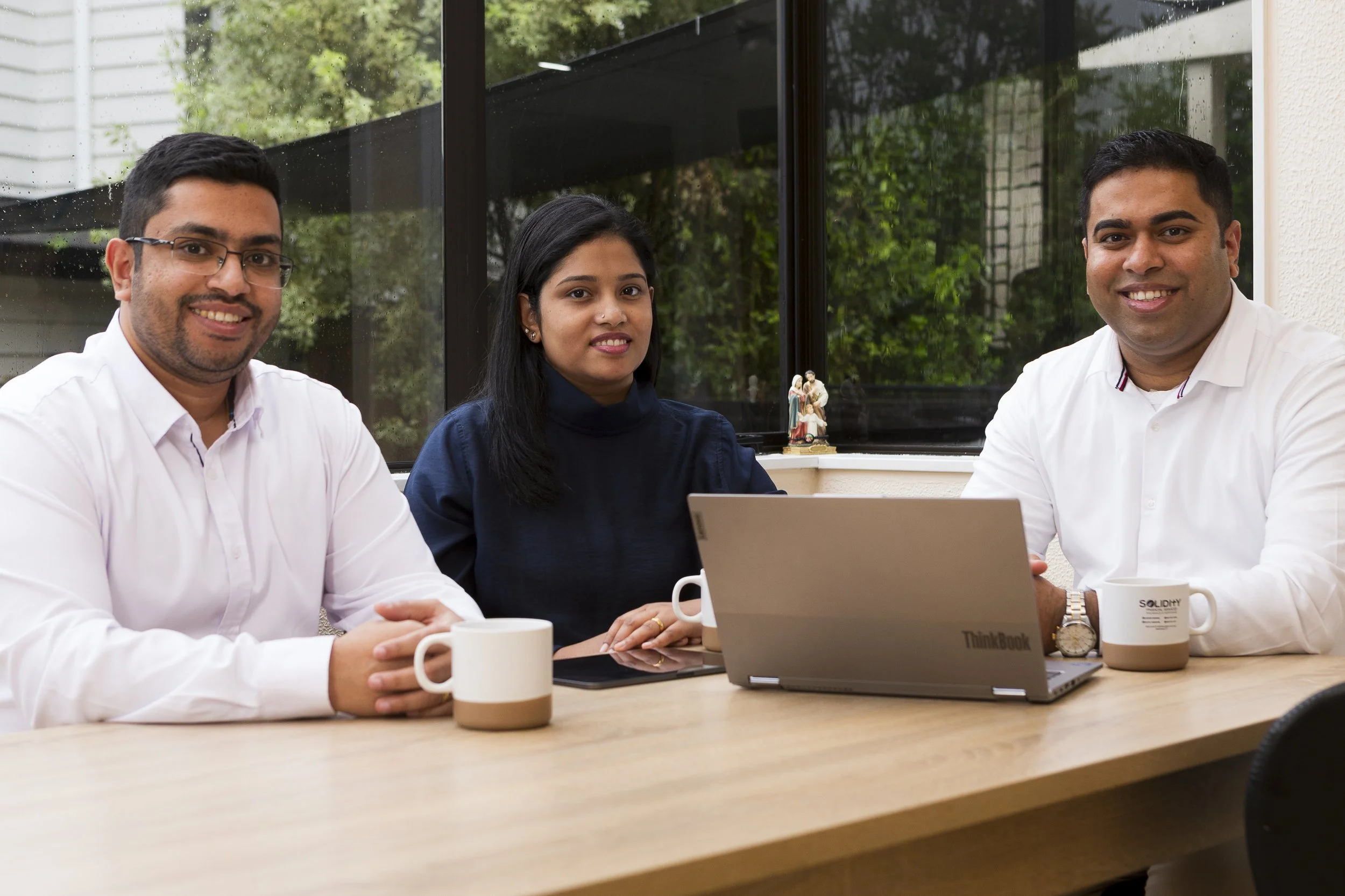 Three people sitting at a table during a meeting or work session, with laptops and mugs on the table, in front of a window with trees outside. Auckland insurance advisors and mortgage advisors.