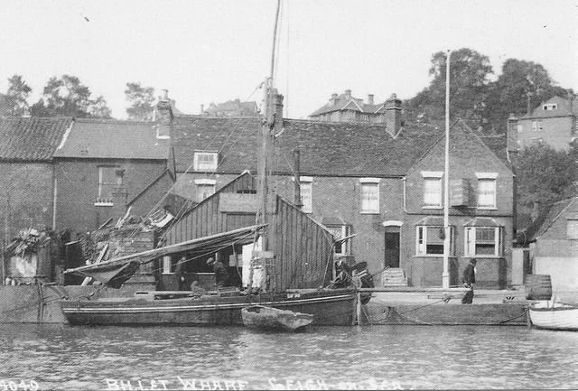 A black and white photograph of a damaged building by the water, with a small boat docked nearby, and a person walking on the dock.