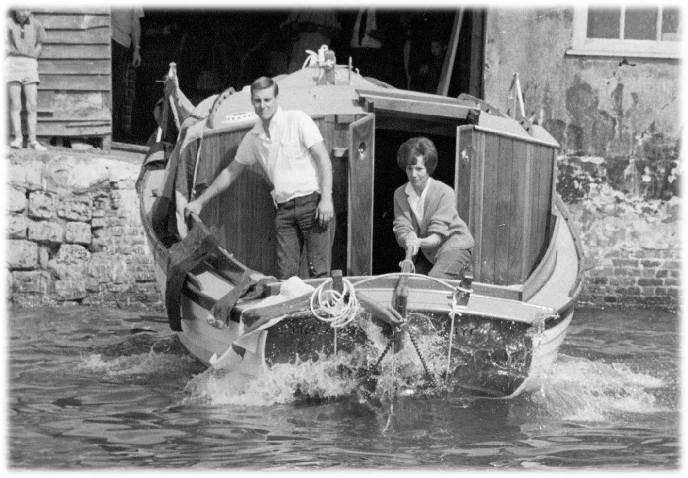 Black and white photo of a small boat with two people on board, a man and a woman, near a stone dock, with buildings in the background.